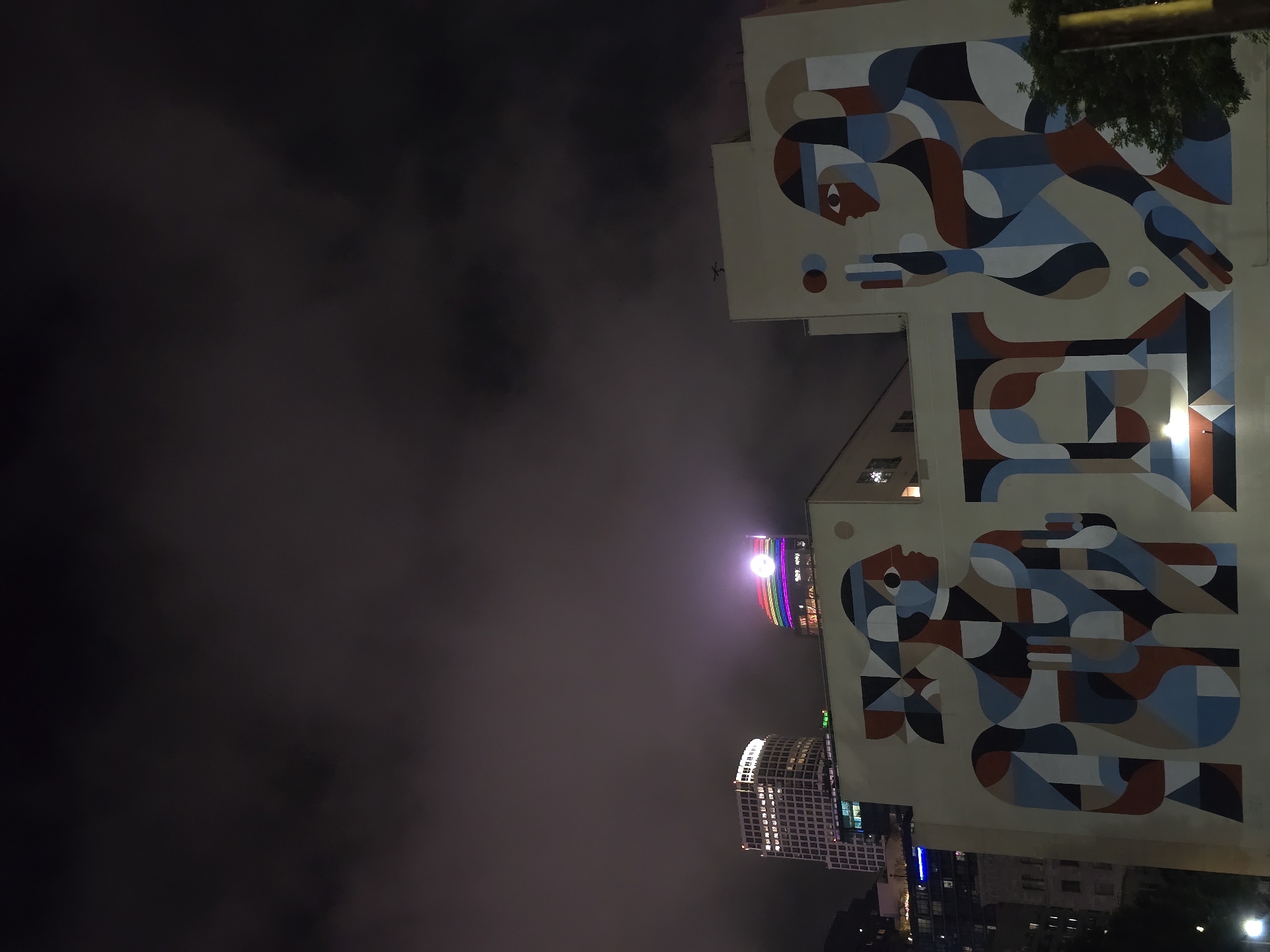 This is a nighttime photograph taken in Los Angeles, California, USA, capturing a mural on the side of a building. The mural features two stylized human figures rendered in a geometric, abstract style with a palette of muted colors including beige, blue, rust, and black. The figures are depicted in profile, facing each other. The figure on the left has dark hair tied back and is holding her hands in front of her. The figure on the right appears to be holding a spherical object and has one hand raised.
Behind the mural, the Los Angeles skyline is visible. Two prominent buildings stand out: a lighter colored, modern skyscraper on the left, and a curved building on the right illuminated with a rainbow-colored light display, suggesting it might be a landmark or a venue celebrating an event. The sky is dark and cloudy, with a diffused glow from the city lights. The overall impression is an urban scene at night, with the vibrant mural contrasting with the muted tones of the night sky and the distant city lights.