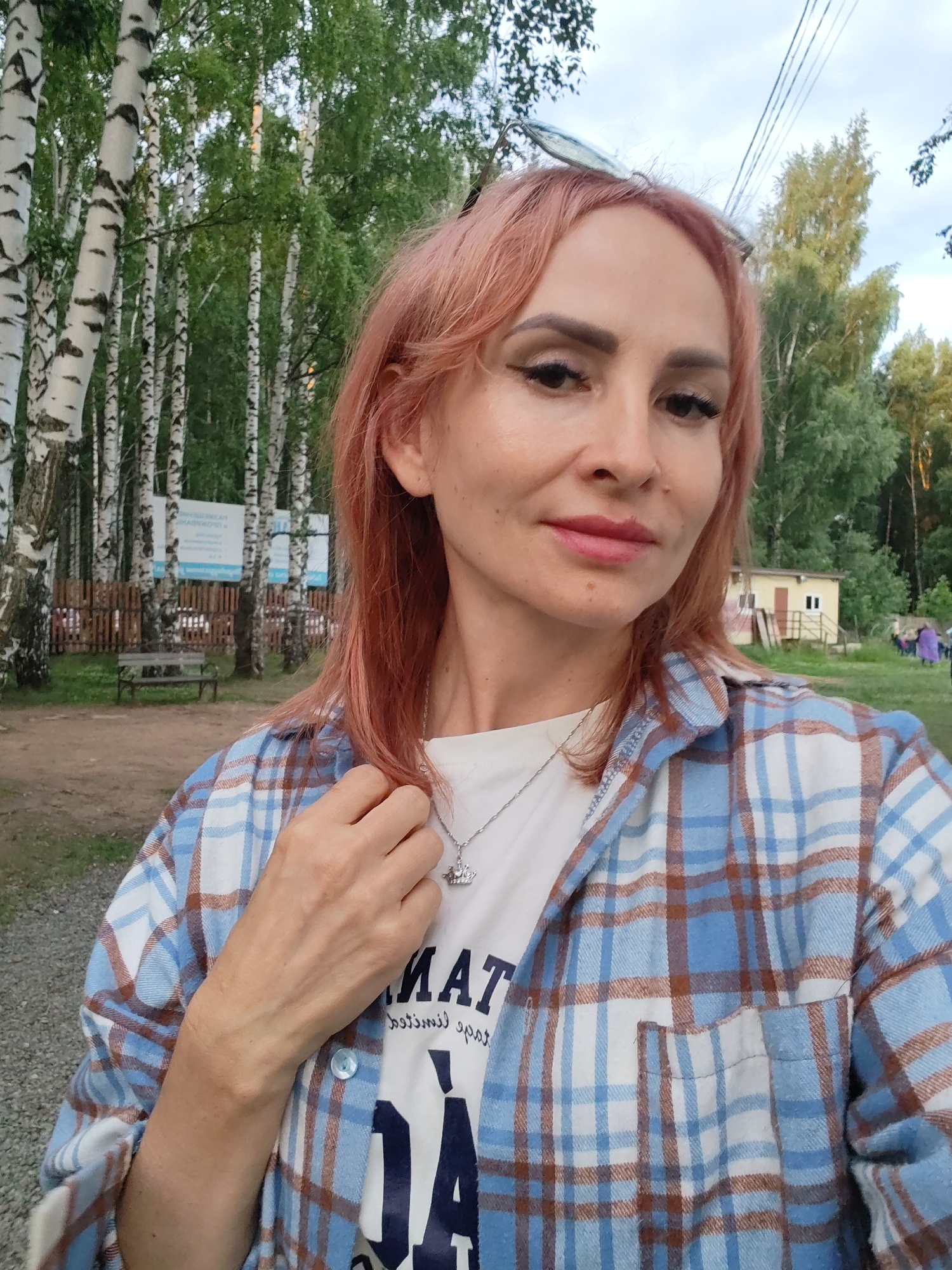 The image is a selfie taken outdoors in Chaikovsky, Russia, with a background of birch trees, a wooden fence, a bench, and a small building. A woman with pink hair is in the foreground, wearing a plaid shirt over a white t-shirt with a logo that is partially visible. She has makeup on and is wearing a necklace with a pendant. Her right hand is resting on her chest. The sky is overcast, suggesting it may be late afternoon or evening. The overall impression is of a casual outdoor setting.