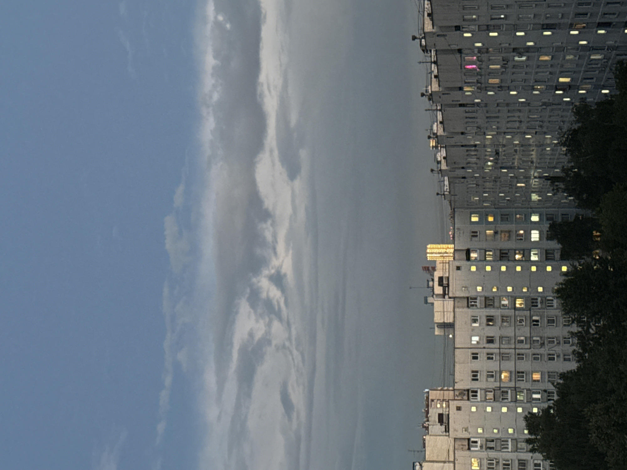 This is a full shot of a multi-story apartment buildings in a city at dusk. The sky is a gradient of light blue to dark gray with scattered clouds. The buildings are a light gray concrete, with many windows lit by yellow light. There are trees in the foreground. In the distance, a taller building with many windows lit by bright yellow lights can be seen.