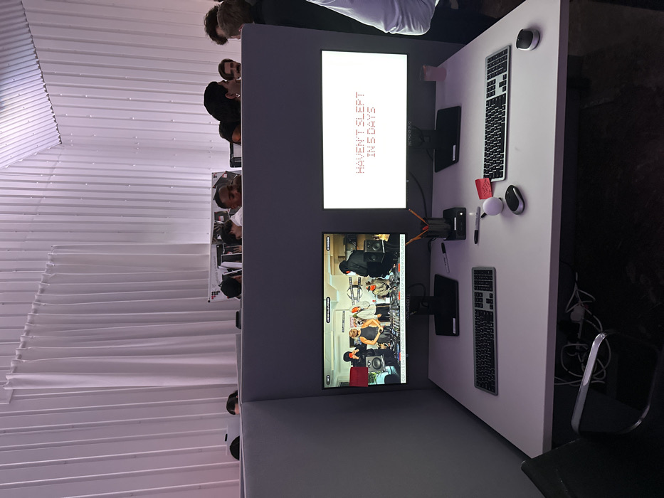 The image depicts an interior scene within an office environment, likely the headquarters of Nothing Technology Limited, situated in the city of London, United Kingdom.
In the foreground, a white desk serves as a workstation, equipped with two black computer monitors on stands. The left monitor displays a live video feed or recording of five individuals in a studio-like setting, featuring audio equipment and one person operating a mixer. Text overlays include "LIVE NOW: (2.30PM EST)", "NOTHING (R) HQ", and "25/25 DAYS OF NOTHING OUTLANDER". The right monitor, set against a dark background, shows prominent white dot-matrix style text reading "HAVEN'T SLEPT IN 5 DAYS", with "NOTHING (R) HQ" visible below.
On the desk, directly in front of the monitors, are two keyboards, a black computer mouse, and a white spherical object. A black pen holder containing pens and pencils is positioned between the monitors. A black Sharpie marker and several red sticky notes, one partially showing "thank you", are also present. A second computer mouse, silver and black, is on the right side of the desk, adjacent to a translucent pink cup. Cables are visible beneath the desk.
The workstation is defined by grey, low-height cubicle dividers. Beyond these dividers, in the background, approximately ten individuals are visible from the shoulders up, suggesting they are either standing or seated at adjacent workstations or mingling. The overall lighting has a cool, purplish tint. The background architecture features corrugated wall panels, partially obscured by white, vertically draped fabric panels extending towards an angled or arched ceiling.