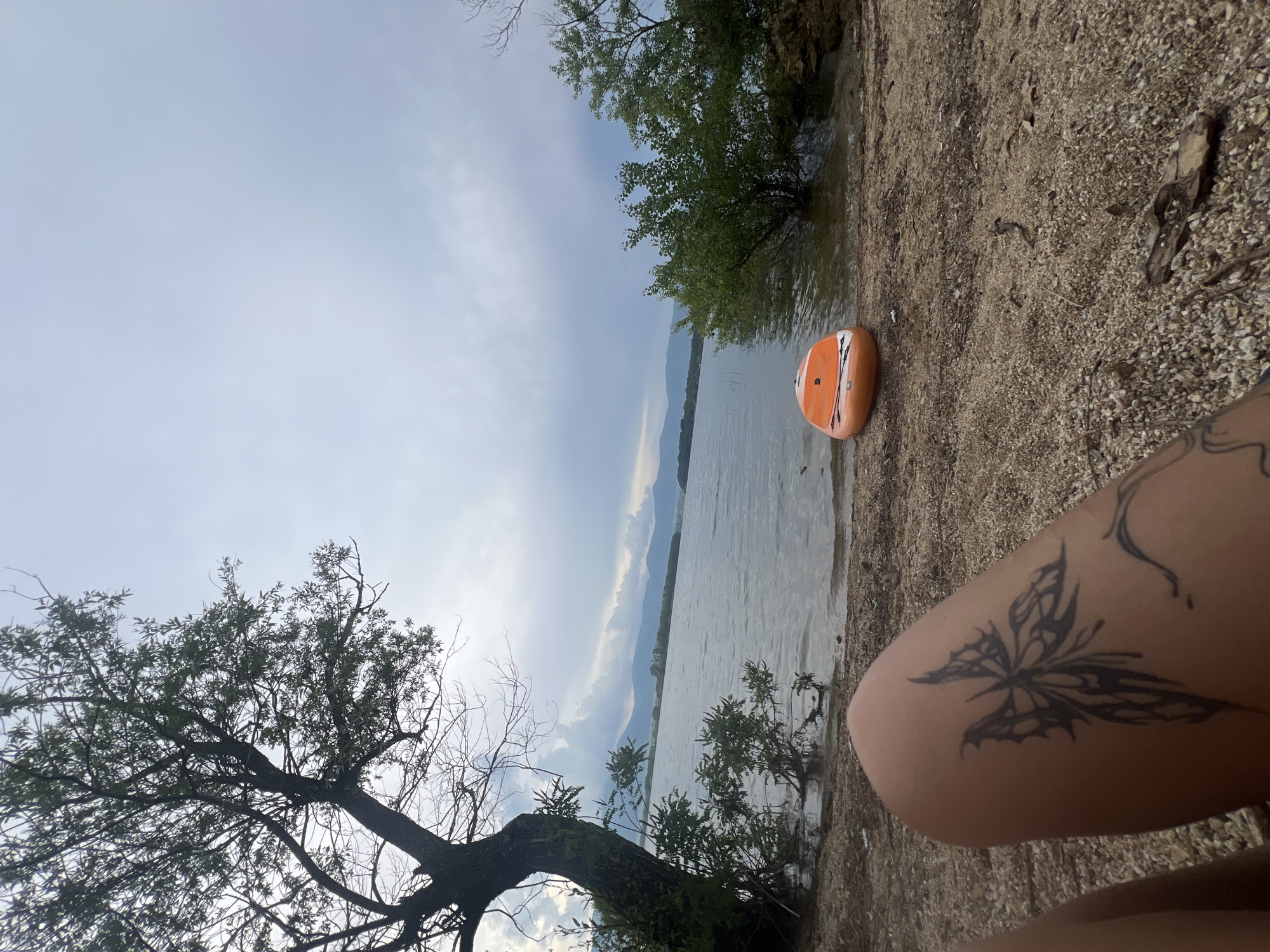 The image depicts a serene lakeside scene at Koprinka Reservoir in Bulgaria. In the foreground, a person's leg with a butterfly tattoo is visible, resting on a sandy shore littered with small stones and debris. The shore leads to the calm water of the reservoir. An orange stand-up paddleboard rests near the water's edge. Across the water, a mountain range is visible under a cloudy sky. Lush green trees and bushes line the shore on the right side of the frame, while a large tree with bare branches stands prominently on the left, near the water. The overall atmosphere is peaceful and natural, suggesting a leisurely day by the lake.