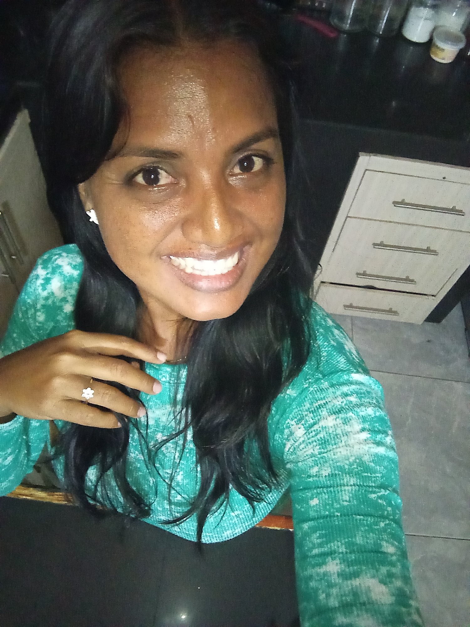 In this image, a woman with dark hair and brown eyes is smiling at the camera. She is wearing a teal and white patterned long-sleeved shirt and a flower-shaped ring on her left hand. A white flower-shaped earring is visible on her left ear. The background is out of focus but appears to be a kitchen, with cabinets and jars visible. The lighting suggests the photo was taken indoors, possibly at night.