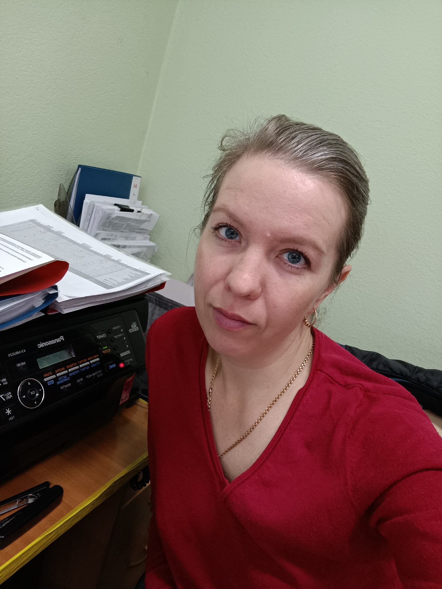 A woman with fair skin, blue eyes, and light brown and grey hair pulled back is featured in a close-up, slightly high-angle shot. She wears a red V-neck long-sleeved top, a gold chain necklace, and small gold hoop earrings. She gazes directly at the camera with a neutral expression.
The setting is an office or workspace, likely in Chaikovsky, Russia. A light green textured wall forms the background. To her left (viewer's right), a black Panasonic KX-MB5050 multifunction printer sits on a wooden desk. A significant stack of paper documents, some in red and blue folders, is piled on and next to the printer. Additional documents and blue binders are visible behind the printer, and a black stapler rests on the desk below it.
The activity suggests a selfie or a casual portrait taken at her workstation.
Visible text on the printer, partially mirrored, includes "Panasonic" and the model number "KX-MB5050". Other legible labels on the control panel, despite being mirrored, include "ОТПРАВКА" (SEND), "КОПИЯ" (COPY), "ФАКС" (FAX), "ПАМЯТЬ" (MEMORY), "НАСТРОЙКИ" (SETTINGS), "ПУСК" (START), "СТОП" (STOP), "OK", "HOT", and "MYK". On a blue folder, "A4" is visible. The documents contain lines of text and numbers, indicative of office paperwork.