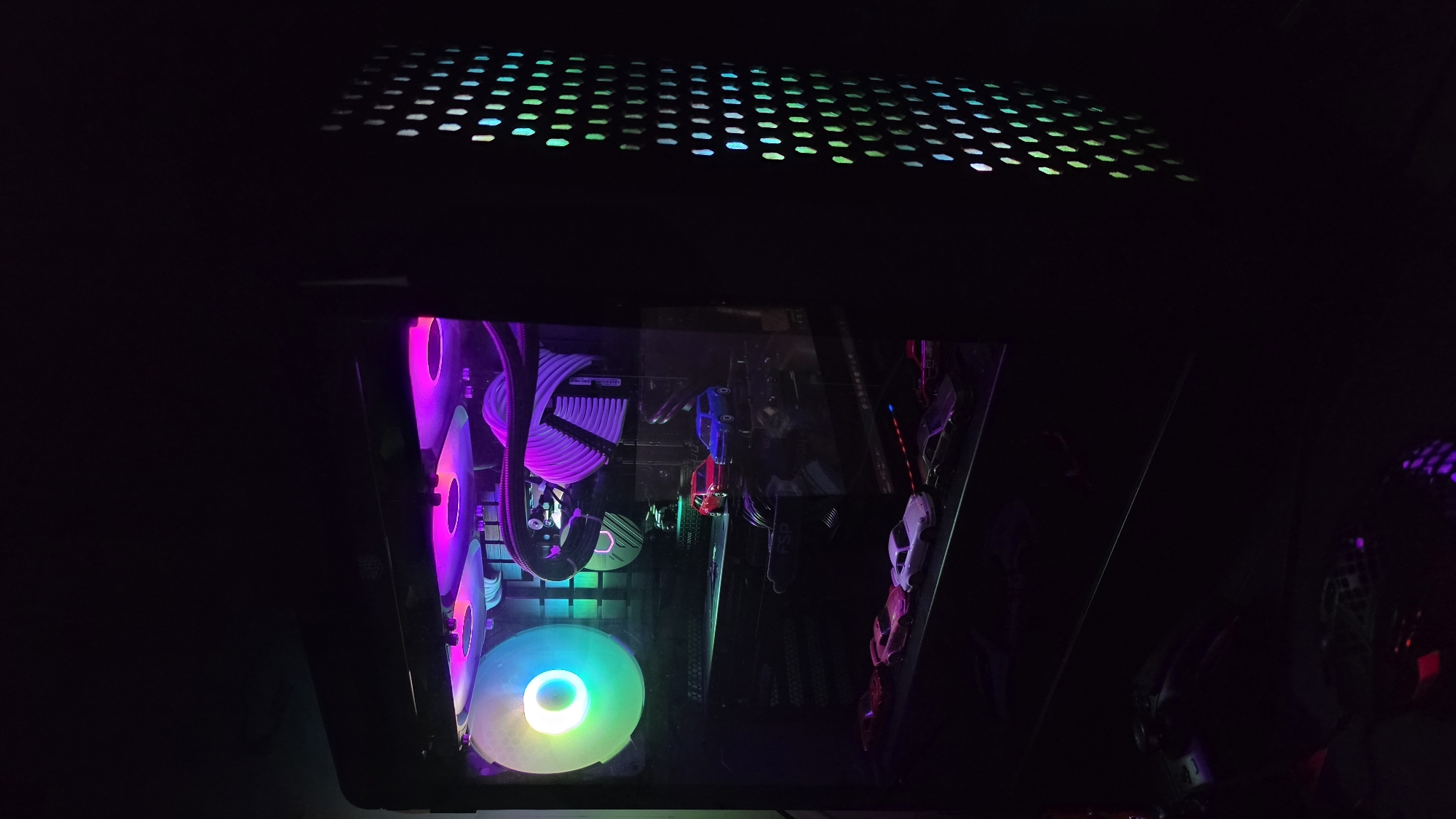 The image shows the inside of a computer tower with RGB lighting. The case is dark, with a perforated top panel allowing light to escape. Inside, there are multiple fans with RGB lighting in various colors, including pink, purple, green, and blue. There are also visible computer components such as a CPU cooler with braided cables and a graphics card. There are toy cars on the right side of the case. The lighting suggests a gaming or high-performance PC setup. There is no indication of the location being Varna, Bulgaria.