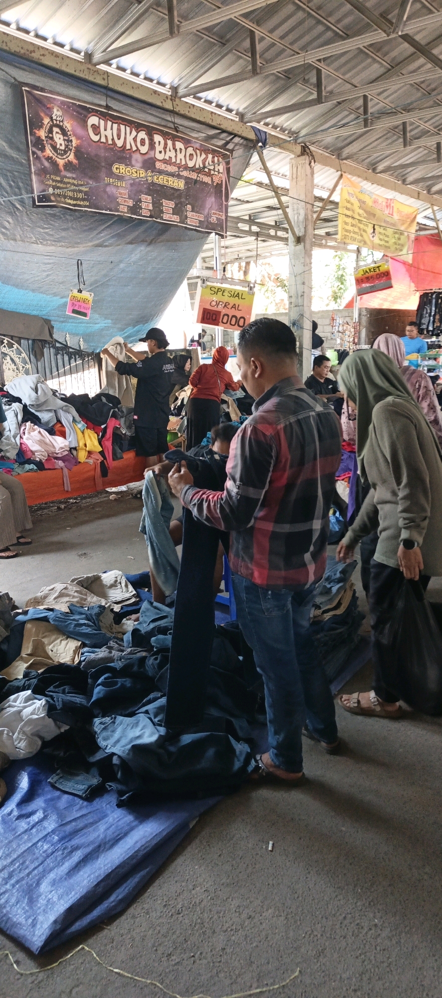 The image captures an outdoor market scene in Jatinom, Indonesia, with a focus on the sale of used clothing. In the foreground, a man in a red and black plaid shirt and blue jeans is examining a pair of dark denim jeans. To his right, a woman wearing a light brown hijab is also looking at clothing. Behind them, a chaotic display of used clothes is spread across tarps and piled high. Several other individuals are visible in the background, some engaged in browsing or selling. Above the stalls, banners are hung, one prominently displaying "CHUKO BAROKAH GROSIR & ECERAN," indicating a wholesale and retail clothing business. Another banner advertises "SPESIAL ORAL 100.000," suggesting a special offer. The environment appears to be a bustling market with a rough, asphalt-like ground surface and a corrugated metal roof overhead, supported by visible metal beams. The overall impression is one of a lively and informal second-hand clothing market.