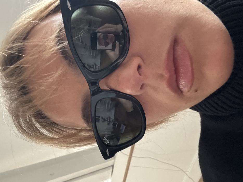 The image is a close-up, low-angle selfie of an individual with light-colored, short-cropped hair and bangs, wearing black Ray-Ban Wayfarer sunglasses and a black ribbed turtleneck sweater. The subject's face is partially visible from the nose down, revealing full, glossy lips and a small dark mole located inferior and lateral to the left corner of the mouth (viewer's left). The reflection in the right sunglass lens (viewer's left) depicts an indoor environment in Paris, France, characterized by a light-colored wall or doorway, and a section of a room containing a dark, indistinct piece of furniture or shelving unit upon which small objects are placed, under what appears to be a framed item or mirror. The reflection in the left sunglass lens (viewer's right) shows the individual holding a dark-colored smartphone, used to capture the image, with a smaller, secondary self-reflection visible on the phone's screen.