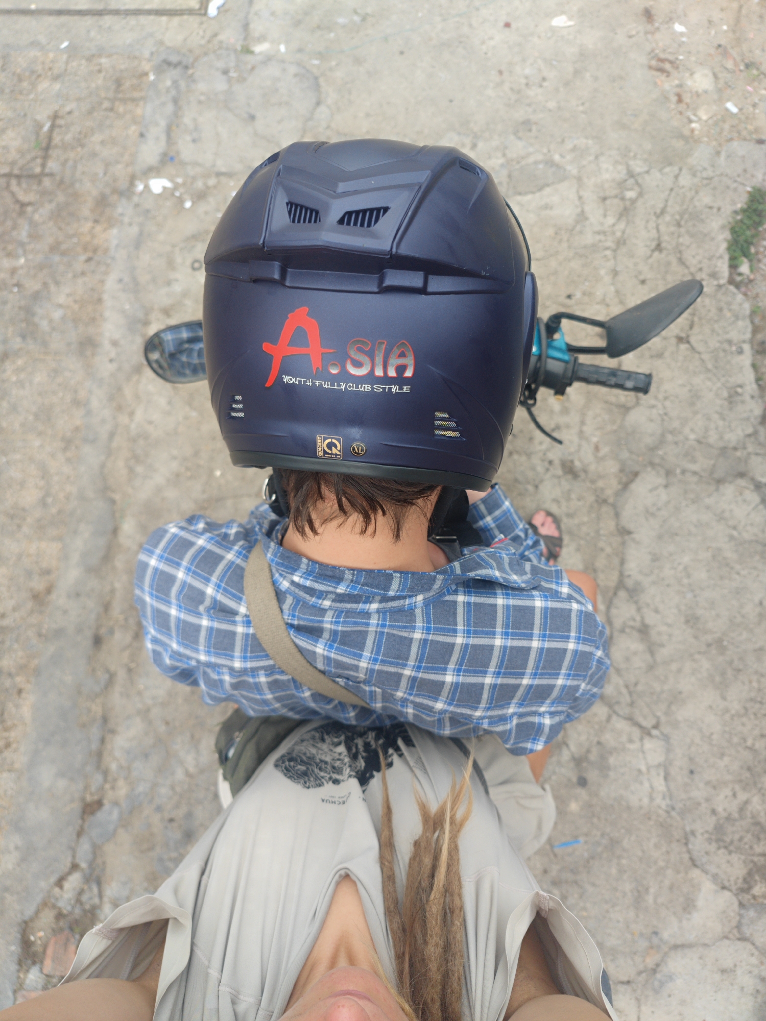 An overhead shot shows two individuals on a scooter in what appears to be Cam Ly, Đà Lạt, Vietnam. The person in the foreground, presumably the driver, wears a blue and white plaid shirt with a light gray strap across their shoulder, indicating they may be carrying a bag. They are also wearing a blue helmet with red and white lettering "A.SIA - YOUTH FULLY CLUB STYLE." The second person sits behind the driver. The concrete ground shows signs of weathering and repair. The scooter is visible, with parts of the handlebars and mirrors in view.
