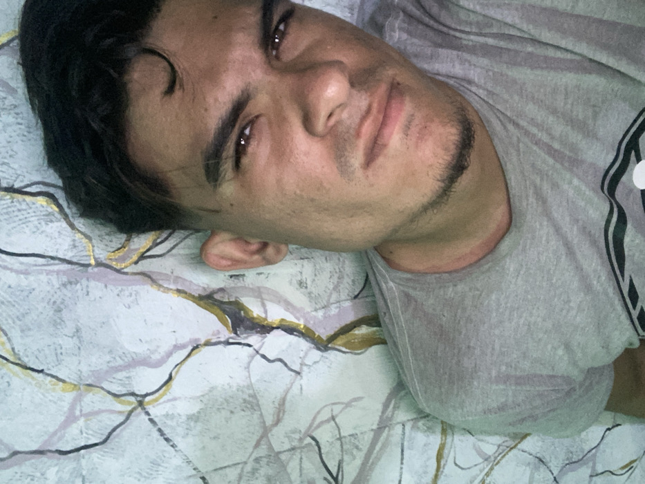 A light-skinned man with dark hair and stubble lies on a patterned bed. He is wearing a gray shirt. The bedspread is white with gold, gray, and dark lines creating a branching pattern. He looks directly at the camera with a slight smirk. His head is resting on the pillow, positioned on the left side of the frame. The shirt covers his right shoulder and upper chest.
