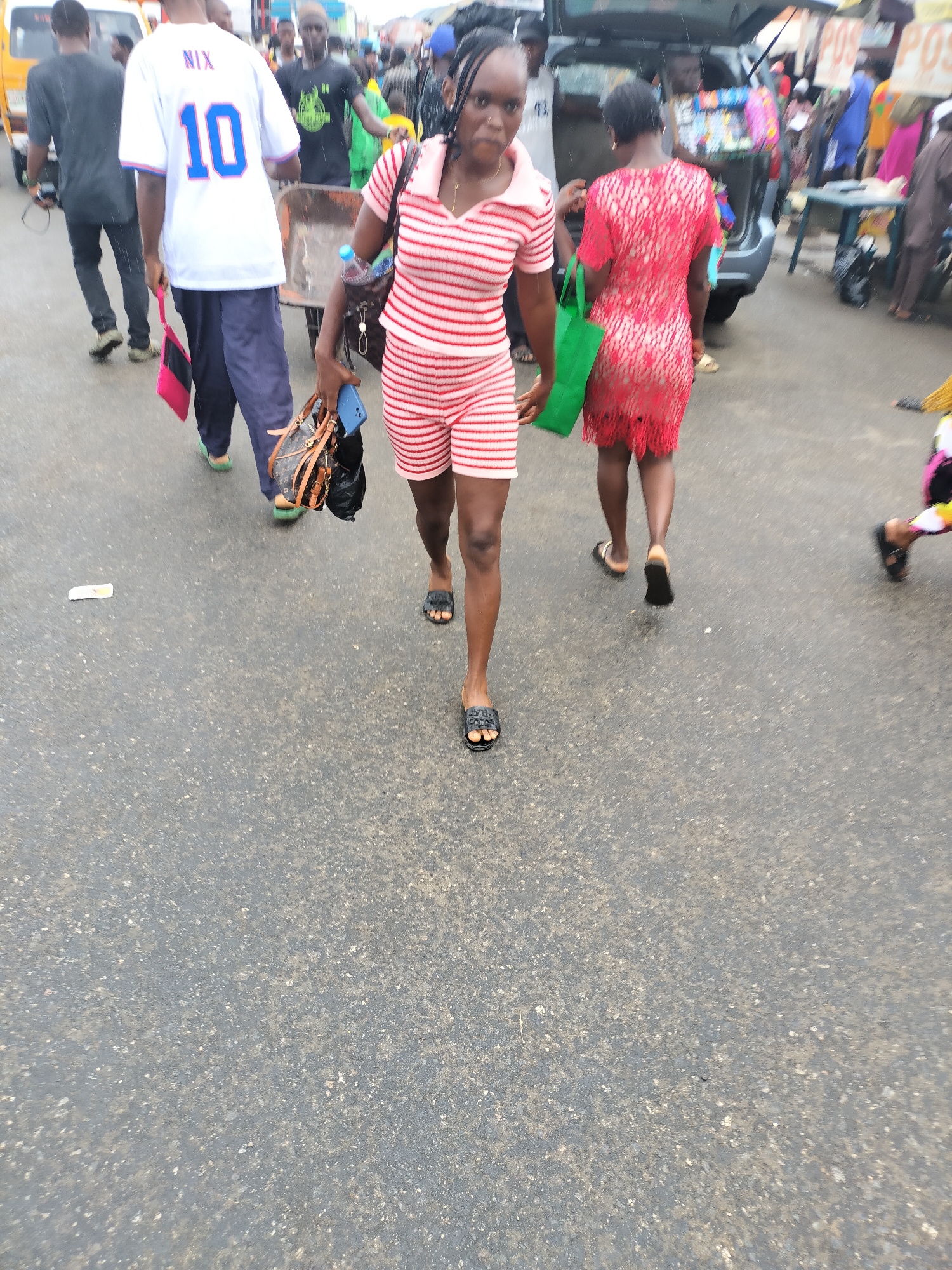 The image captures a street scene in Ikorodu, Nigeria. The focus is on a woman walking towards the camera, wearing a red and white striped polo shirt and shorts. She carries a handbag and a green shopping bag. Behind her, a woman in a red dress walks in the same direction. To the left, a man in a white jersey with the number "10" walks alongside another man in a dark shirt, both moving away from the camera. In the background, there is a mix of pedestrians and vehicles. A car with its trunk open is visible, with people seemingly loading or unloading items. The environment appears to be a busy market area, possibly after or during rainfall given the wet asphalt road. There are various indistinct objects and structures in the background, suggesting a bustling urban setting.