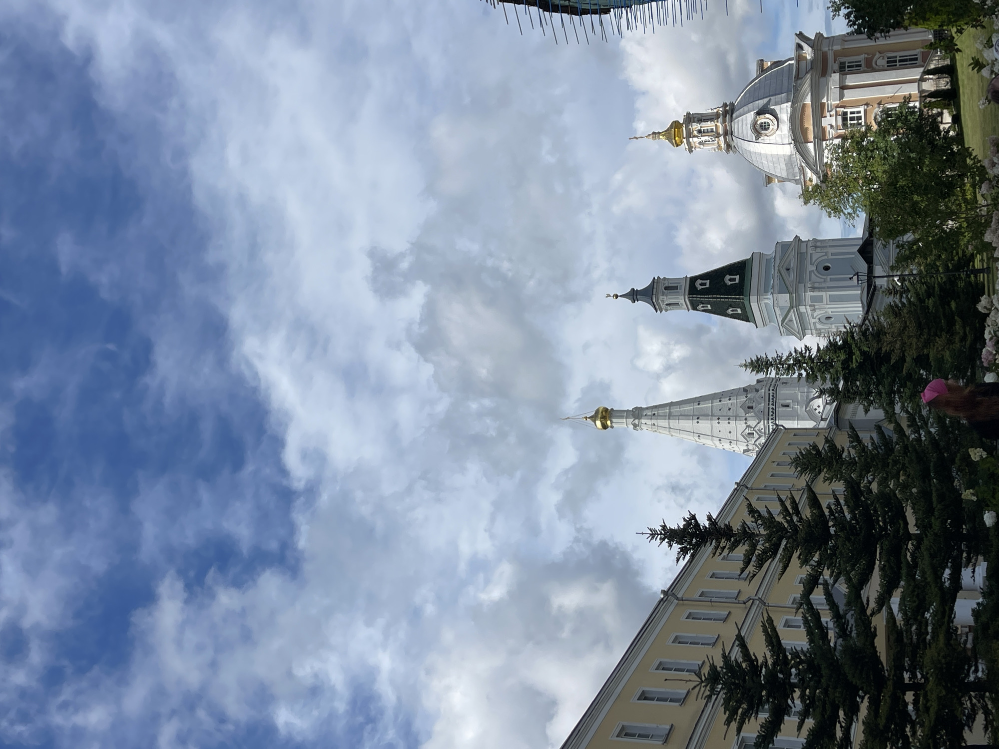 A medium-shot, eye-level vertical photograph depicts a portion of the Holy Trinity-St. Sergius Lavra complex in Sergiyev Posad, Russia. The upper half of the image is dominated by a dramatic sky filled with fluffy white clouds against a bright blue background. In the lower half, a pale yellow building with white trim and numerous windows runs along the left side of the frame. To the right of this building, several tall, dark green coniferous trees are visible.
Behind the trees and the yellow building, the iconic architecture of the Lavra is prominent. A tall, white bell tower with a pointed, white spire topped by a golden dome rises towards the sky. To its right, a second, more elaborate structure features a large, silver dome, also topped with a golden cross. This dome is part of a pinkish-orange building with white architectural details and windows. Further to the right, the edge of another dark, angular structure, possibly under construction or restoration, is visible. In the foreground, at the bottom edge of the frame, the back of a person wearing a pink hat is visible, facing away from the camera and towards the Lavra structures. The overall scene suggests a bright, possibly breezy day.