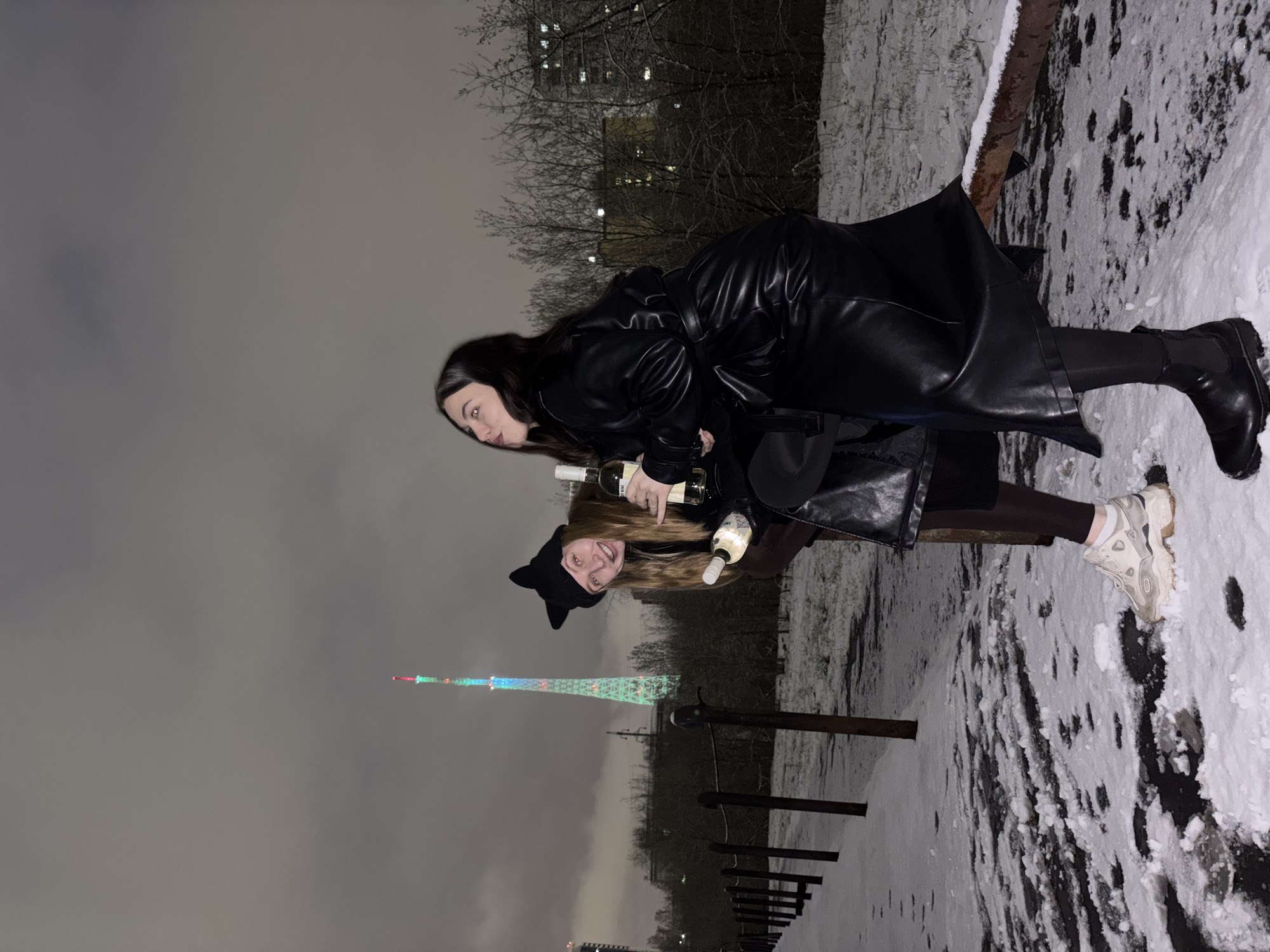 The image depicts two young women outdoors at night in a snowy, winter setting, possibly a park or pathways. The original image is rotated 90 degrees clockwise, but it is described as if correctly oriented in portrait.
The woman in the foreground on the left wears a long black, shiny, leather-like trench coat and dark, chunky-soled boots over dark tights. She has long dark hair and a neutral to slightly pouty expression as she looks towards the viewer. She is holding two bottles of white wine.
Behind her and slightly to the right, another woman is visible. She has lighter, possibly brown or blonde, hair and wears a black knit hat with prominent cat ears. She is smiling broadly, showing joy, and is also holding two bottles of white wine. She wears lighter-colored sneakers. Both women are leaning against a low, dark railing or bench, which is partially covered in snow.
The ground is heavily covered in patches of snow and dark, wet earth. Bare tree branches are visible in the background against a dark, overcast night sky, with faint lights of buildings in the distance. A prominent feature in the background is a tall, slender telecommunication tower, which is illuminated with multicolored lights (red, blue, green). Given the context of Perm, Russia, this is likely the Perm TV Tower.
The overall atmosphere suggests a cold, festive evening event, with the women enjoying themselves despite the winter weather. No discernible text is visible on the bottles or elsewhere in the image.
