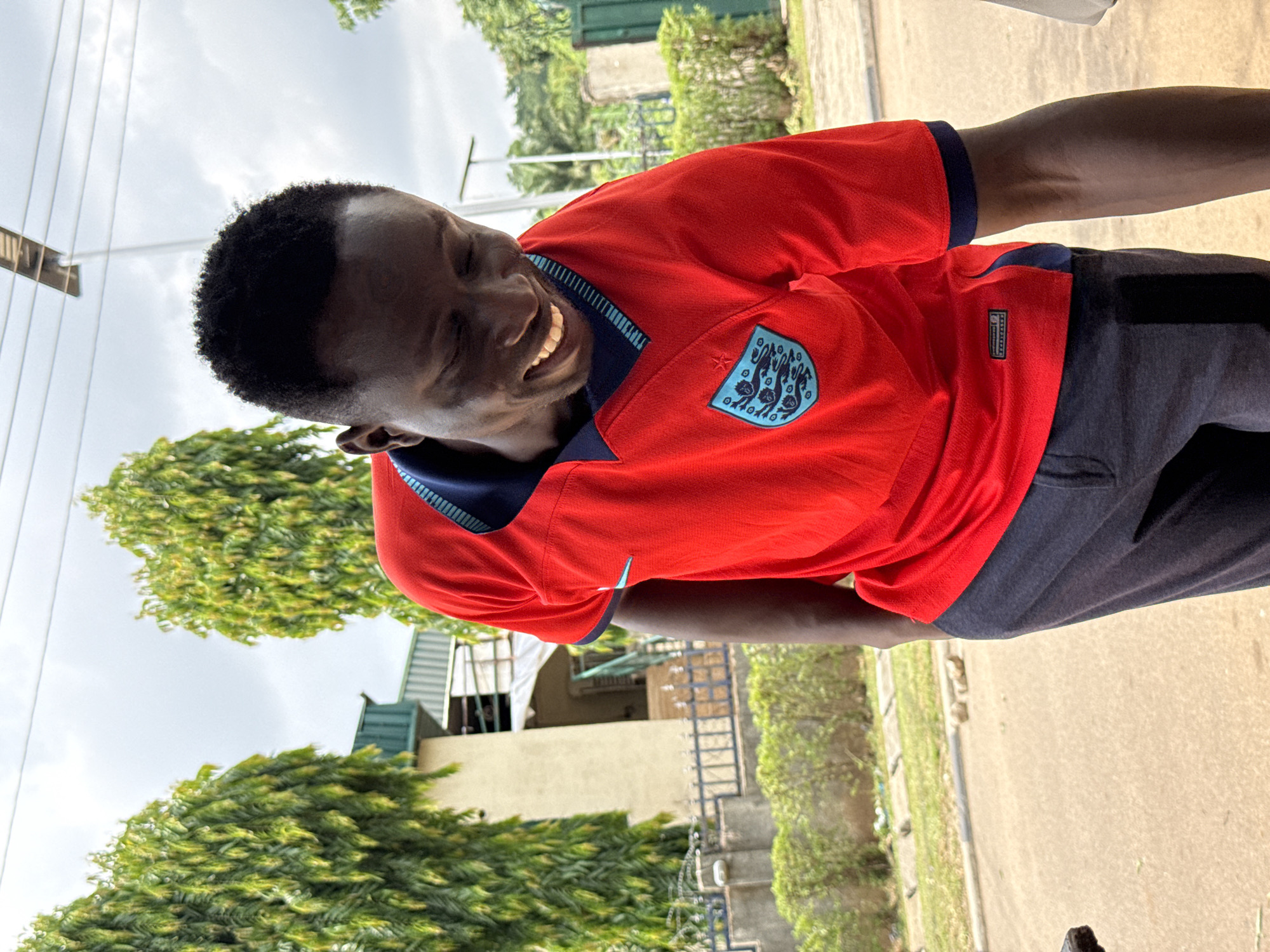 The media file shows a young Black man from the chest up, smiling broadly and looking to his left. He is wearing a bright red short-sleeved shirt with a navy blue collar and trim. On the left side of his chest, there is a light blue crest with a white outline and a white star above it. The shirt appears to be a soccer jersey. He is also wearing dark gray pants or shorts. The man's expression suggests happiness or amusement.
The background is slightly out of focus but provides context. To the left of the man, there are green trees with dense foliage, and beyond that, a cloudy sky. To the right, there is a paved surface that appears to be a road or pathway, with more greenery and some buildings visible in the distance. The overall lighting suggests it is daytime, possibly late morning or afternoon, with the sun casting some shadows. The presence of lush greenery and the style of buildings hint at a warm climate, consistent with the provided location context of Abuja, Nigeria. There are also some electrical wires visible in the upper left corner of the image. A small white tag with black text is visible on the lower right side of the man's shirt, below the crest. The image is oriented vertically.