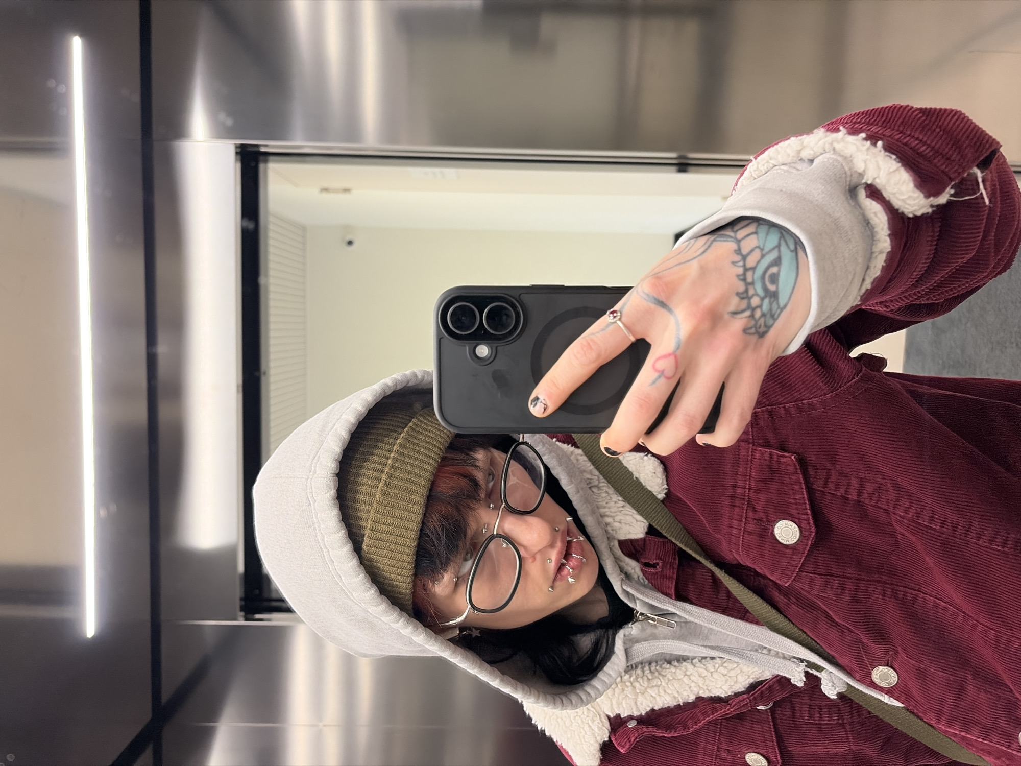 A person is taking a selfie in what appears to be an elevator. The person is wearing a green beanie, a grey hoodie with a faux fur lining, and a burgundy corduroy jacket. They have numerous piercings on their face and a tattoo on their hand. The person is holding a black smartphone with two cameras. The background shows a mirrored surface with a vertical light fixture. The reflected image shows a white interior space. There is no specific visual information in the image to confirm the location in Busmantsi, Bulgaria.