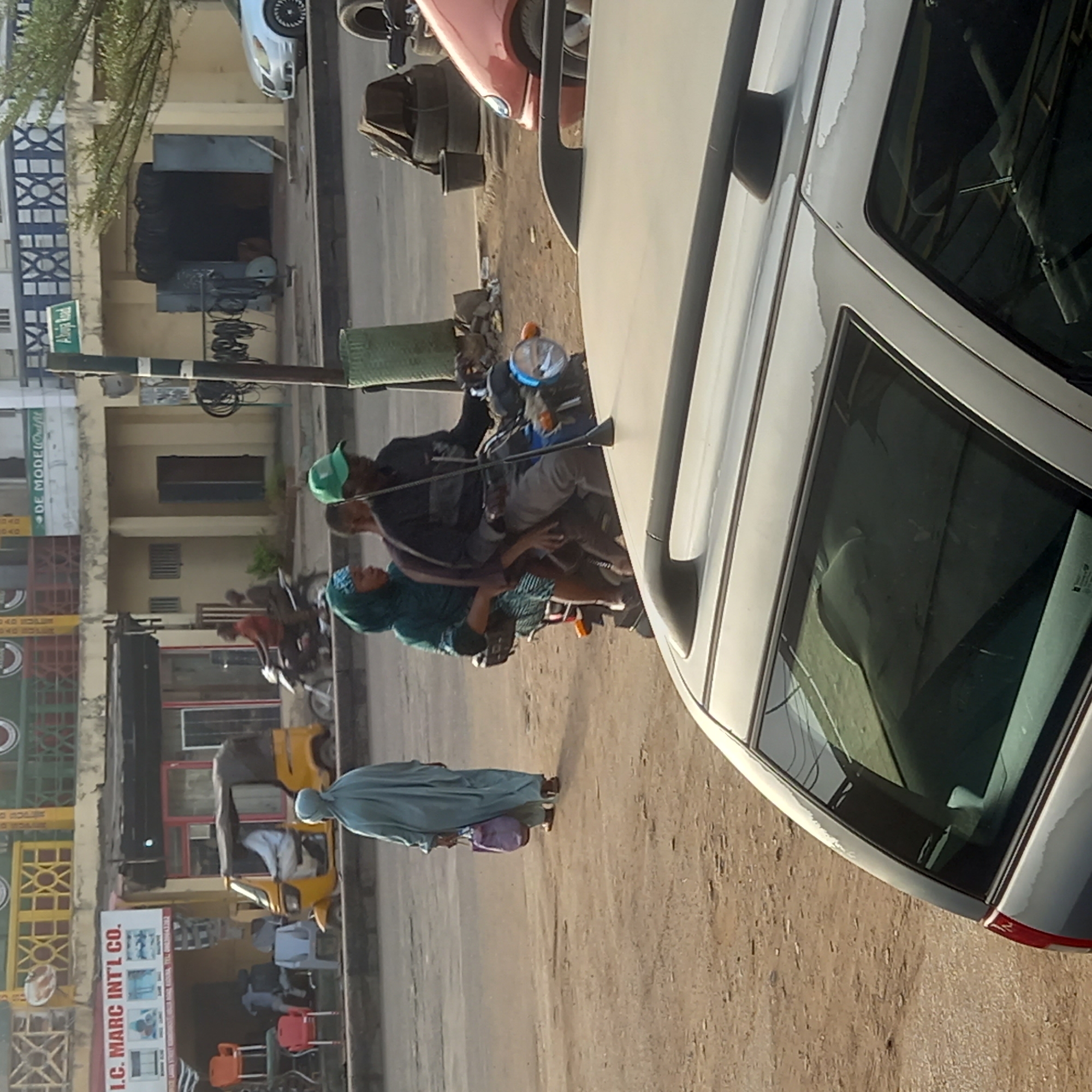 The image depicts a bustling street scene in Dadi Riba, Nigeria, bathed in bright daylight. The view is slightly angled, looking down a road bordered by buildings and commercial spaces.
In the foreground, a silver car is parked on the right, its roof showing noticeable paint peeling. The ground beside the road is unpaved and appears dusty. A utility pole with multiple wires stands centrally, separating the parked car from the moving traffic and pedestrians.
Several people are visible. Two individuals are on a motorcycle: the driver wears a green cap, and the passenger, a woman, is adorned with a teal head covering. Further down the street, another person in a red shirt is seen on a motorcycle. A woman, dressed in a light blue or teal abaya and carrying a purple shoulder bag, walks along the unpaved path on the left, moving away from the viewer. A yellow Keke NAPEP (tricycle taxi) is parked near a storefront, with a person visible inside.
The buildings lining the street house various businesses. Visible signage includes a green sign partially reading "Alhaj...", a white sign stating "DE MODEL AND OUTFIT", and another prominent sign for "I.C. MARC INT'L CO.". Commercial items like stacked tires and an assortment of chairs are arranged outside some establishments, indicating active commerce. The buildings feature distinct patterned grilles or fences.
The scene portrays a typical busy day in an urban or semi-urban setting, characterized by local transportation, commercial activity, and everyday life. No particular emotions are discernible from the individuals.