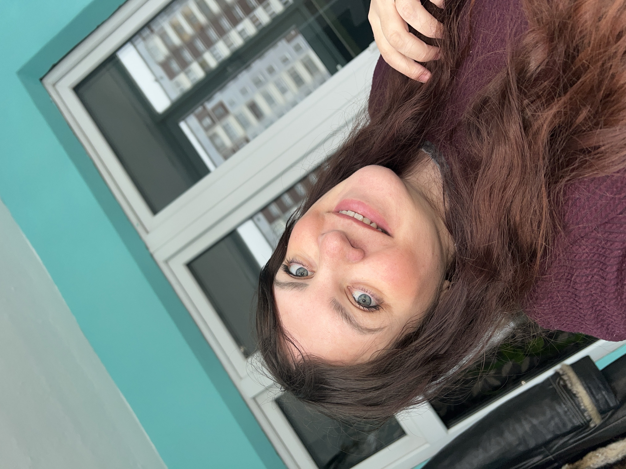 This image is a rotated, close-up portrait of a woman, likely a selfie. She has long, wavy, reddish-brown hair and striking light-colored eyes. The woman is looking directly at the camera with a subtle, pleasant smile, her lips slightly parted to reveal a hint of her teeth. She has a fair complexion and appears to be wearing a dark reddish-purple knitted garment. Her left hand is partially visible at the top of her head, gently touching her hair.
The background features a large window with a white frame, appearing at an angle due to the image's rotation. Through or reflected in the window, a multi-story building with numerous windows is visible, suggesting an urban setting. To the left of the woman, a prominent turquoise-colored wall creates a vibrant contrast with a lighter wall or surface below it. In the bottom right corner, a dark, textured object, possibly made of leather with a fur or rough fabric trim, is partially visible.
The lighting appears soft and natural, consistent with daytime. The woman's expression conveys a sense of calm and approachability. There is no visible text in the image. The composition and angle give the portrait an informal and personal feel.