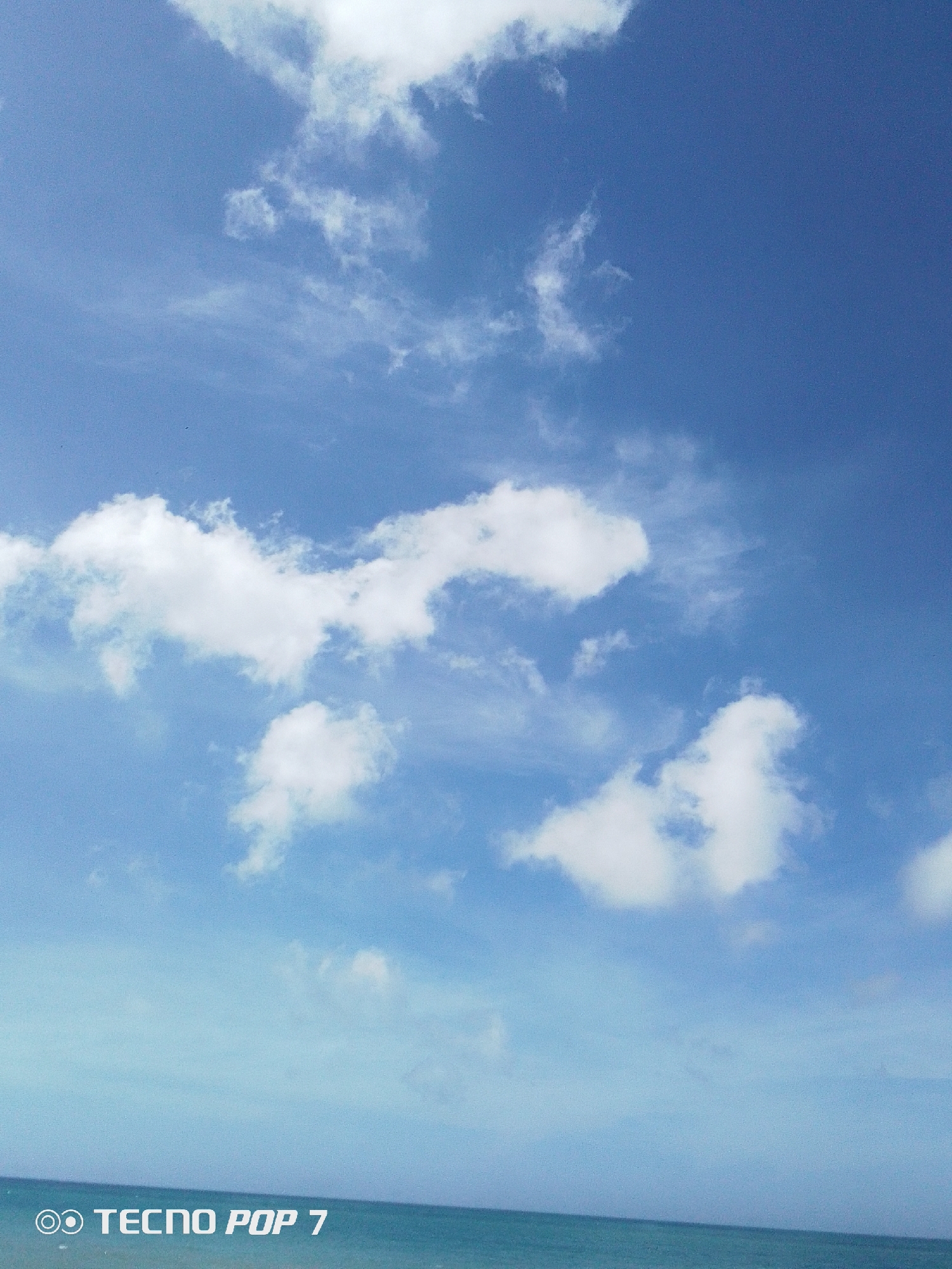 The image depicts a daytime scene in Pedro González, Venezuela, featuring a blue sky with scattered cumulus clouds and a visible horizon line indicating the ocean. The sky occupies the majority of the frame, showcasing varying shades of blue. The clouds are white and fluffy, with different sizes and shapes. The ocean appears as a narrow strip at the bottom of the image, in a slightly lighter shade of blue compared to the sky. The text "TECNO POP 7" is visible in the lower left corner, indicating the device used to capture the photo.