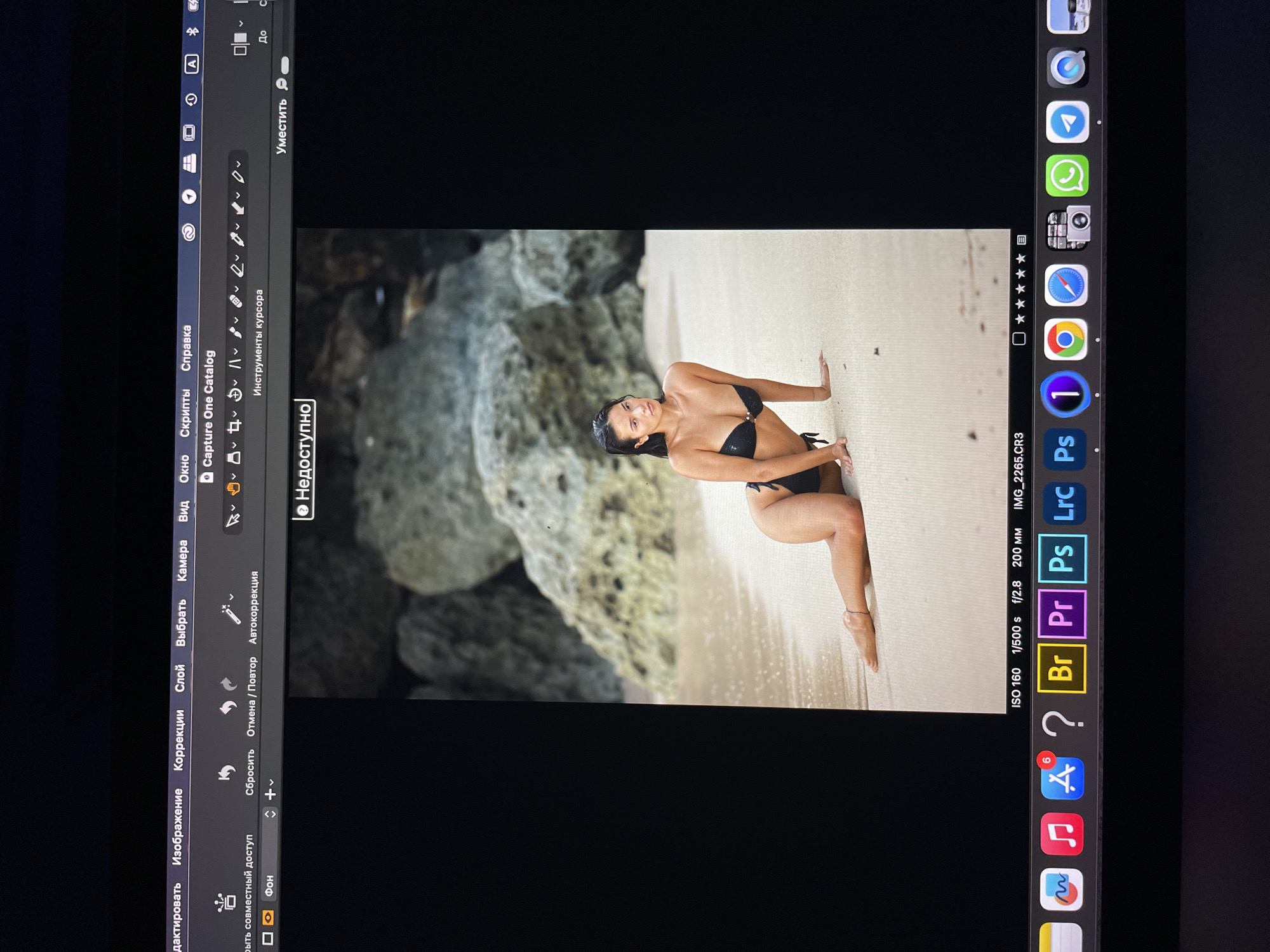 The image shows a woman in a black bikini posing on a sandy beach. Large rocks are visible behind her. The photo appears to be open in an image editing program on a computer screen. The software interface and settings are visible, including menu options like "Image," "Correction," and "Layer." The bottom of the screen displays icons for several applications, including Adobe Photoshop, Lightroom, and Capture One. The camera settings for the photo are shown: ISO 160, 1/500 s, f/2.8, 200 mm. The location is not identifiable from the image.