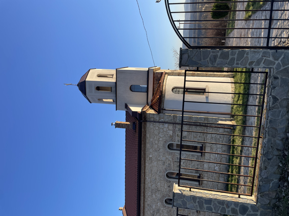 The image displays a church building situated in Cherni Vrah, Bulgaria, under a clear, bright blue sky. The main structure of the church features exterior walls constructed from irregular, light-colored stone masonry and is topped with a red-brown tiled roof. Several arched windows with dark frames are visible along the stone wall. A prominent, light-colored bell tower, potentially stuccoed white, rises from the front-right section of the church, capped with a dark, octagonal-shaped roof that culminates in a golden cross. The tower includes arched openings near its summit and a rectangular window positioned lower. A rounded, white-stuccoed apse or annex, also featuring an arched window, is attached to the base of the tower. A brick chimney with a metal cap is visible on the main roofline.
In the immediate foreground, a black metal fence composed of vertical bars sits upon a base of irregularly cut stone, matching the church's stonework. To the right, an open, arched black metal gate, supported by stone posts, reveals a paved pathway descending into the distance. Green grass covers the ground within the fenced perimeter. Beyond the gate and path, a panoramic view of distant hills or mountains is visible under the clear sky, indicating an elevated position. The scene is illuminated by strong natural sunlight, casting distinct shadows. No individuals or active interactions are present.