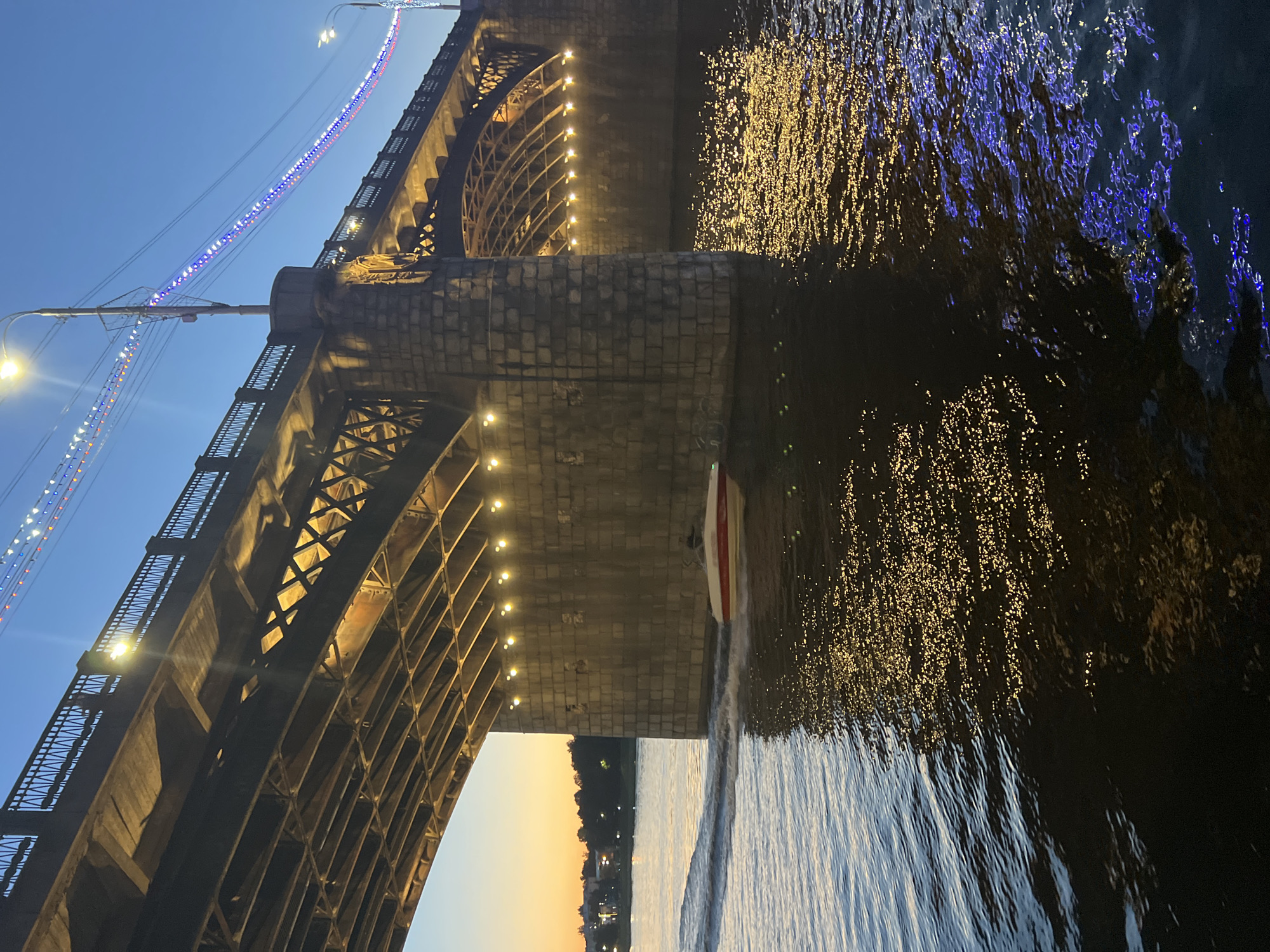 The image depicts a motorboat moving through the dark water under a large, illuminated bridge. The bridge is constructed of stone pylons and steel supports, with lights strung along its railings and under its structure, casting a warm glow on the water. The water reflects these lights, creating shimmering patterns of yellow and blue. The sky above is a deep twilight blue, with a hint of the sunset's orange and pink visible on the horizon in the distance, silhouetting trees and buildings. The motorboat, white with a red stripe, is leaving a wake as it travels from left to right across the frame. This scene is captured on the Volga River in the city of Tver, Russia, near the Starovolzhsky Bridge.