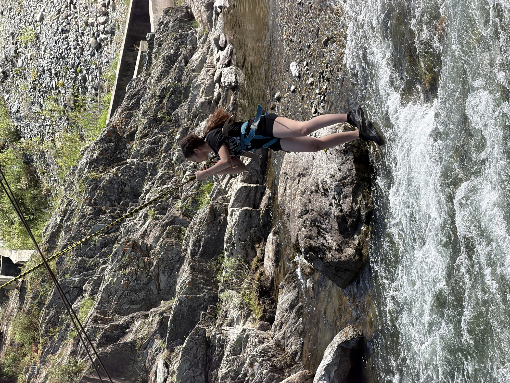 A young female, wearing a black t-shirt, black shorts, and a blue harness, is standing on rocks in a shallow, rocky river. Her hair is dark brown and pulled back in a ponytail. She is holding onto a yellow and black rope that is attached to cables overhead. The river has fast-moving water. In the background, there is a rocky, sloped hillside covered with sparse vegetation and trees. The location is identified as Chemal, Russia.