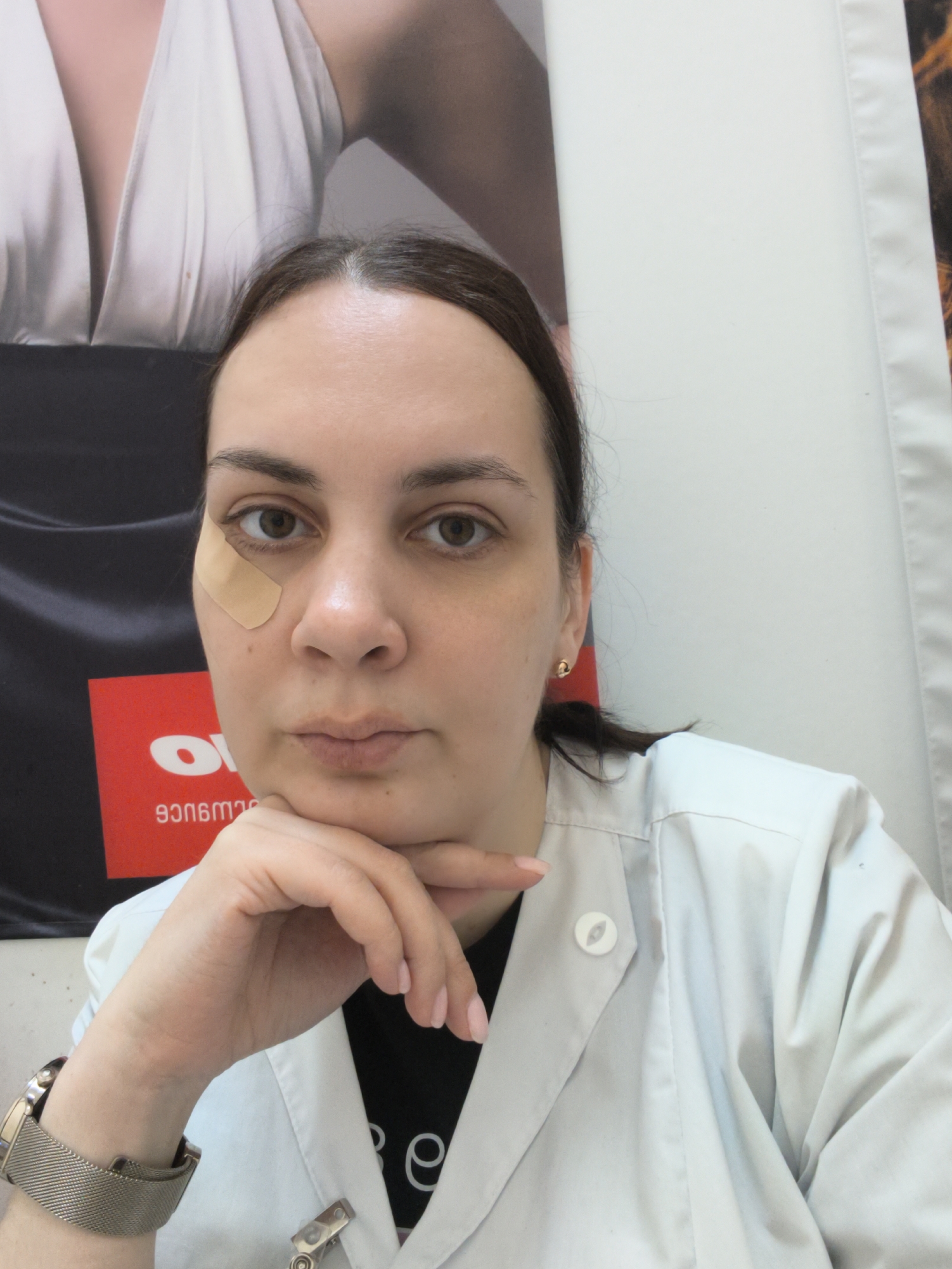 The image shows a woman in a close-up, chest-up indoor selfie. She has dark hair, dark eyes, and a neutral, direct gaze, with her chin resting on her right hand. A small, flesh-toned bandage is visible below her left eye. She is wearing a white lab coat or professional jacket over a black t-shirt, with some partial text, possibly "e," visible on the t-shirt. On her left wrist, she wears a gold-toned watch with a metallic mesh band. A small, circular white pin with an oval design is attached to the lapel of her white coat. A single gold stud earring is visible in her right ear. Her nails are manicured with light pink polish.
The background consists of various large posters or banners. To the left, a black banner features a red rectangular section with reversed white text in Georgian script, reading "პრომო" (promo) and "ფასდაკლებით" (discount). Above this, a portion of another poster displays a person wearing a white, draped garment with a deep neckline. To the right, a plain white panel, possibly a wall or another banner, is visible. The setting appears to be an indoor commercial or professional environment, potentially a clinic, office, or shop, indicated by the white coat and promotional materials. The lighting suggests an artificial indoor source.