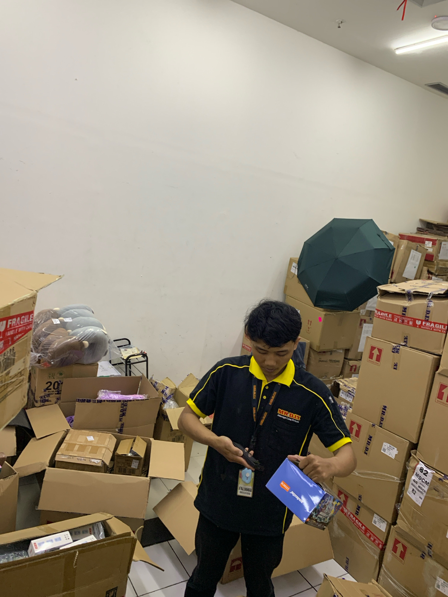 In a warehouse setting in Malang, Indonesia, a young man wearing a black and yellow MR.DIY polo shirt is holding a blue box. The warehouse is filled with stacks of cardboard boxes, some of which are labeled "FRAGILE." To the left of the man, a dark green umbrella is open and leaning against a stack of boxes. The floor is tiled, and the walls are white. The lighting appears to be from overhead fluorescent lights. The man's expression is focused as he examines the blue box, which has graphics on it.