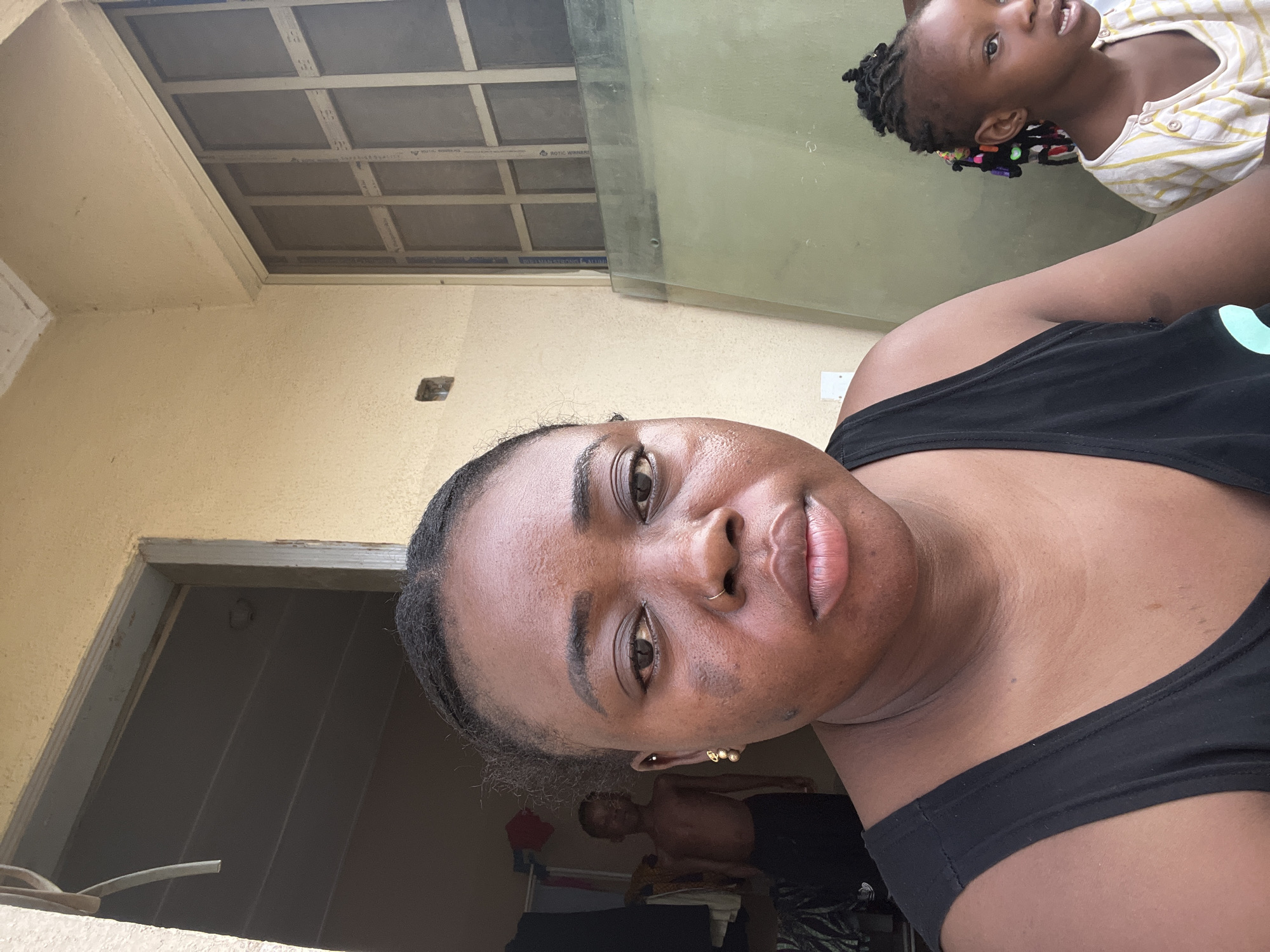 A woman with dark hair, wearing a black top, is pictured in a close-up selfie taken indoors. Behind her, a young girl with braided hair is partially visible near a window. In the background, through a doorway, a shirtless man is seen. The environment suggests a residential interior with painted walls, a ceiling, and an open doorway. The location is Madalla, Nigeria.