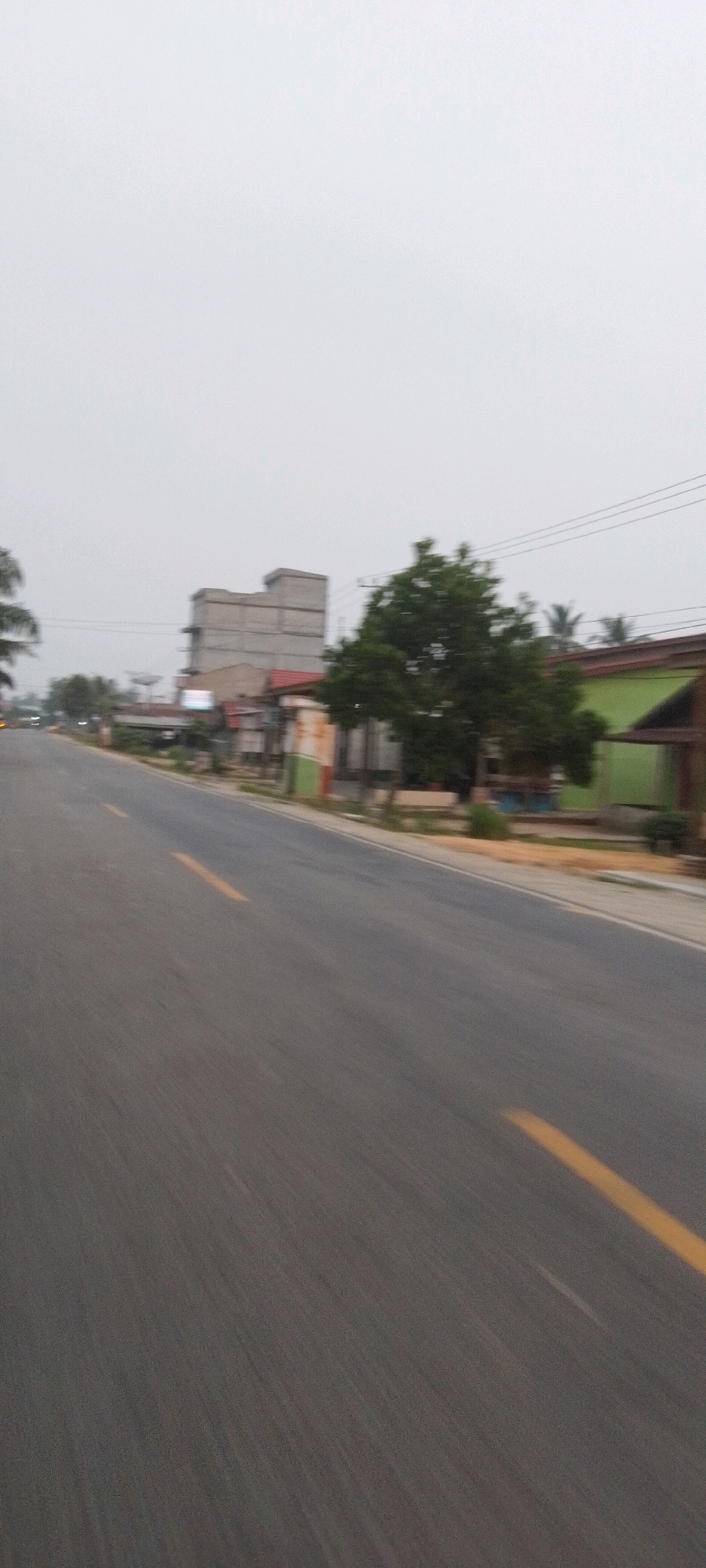 The image depicts a road with a yellow line marking the lane, indicating a street in Teluk Pulau Hulu, Indonesia. The road is paved and appears to be in moderate condition, with some signs of wear and tear. The surrounding environment consists of a few buildings and trees, suggesting a somewhat rural or suburban setting. The sky is overcast, creating a soft, diffused light. The overall impression is that of a typical street scene in Indonesia, possibly during a cloudy day. The image appears to have been taken from a moving vehicle, as evidenced by the motion blur.