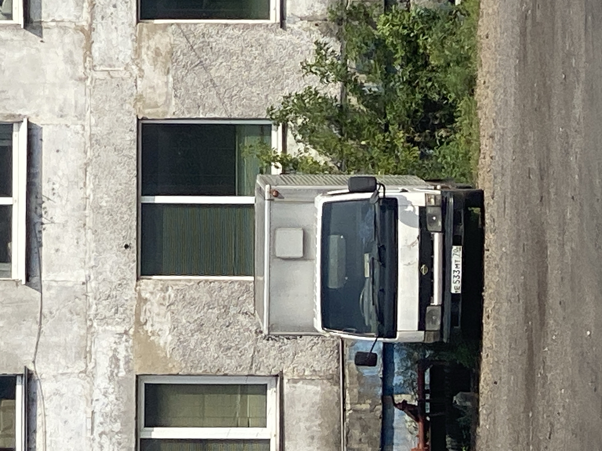 The image shows a white box truck parked on a gravelly surface in front of a worn, concrete building. The building has at least two windows with blinds. To the right of the truck, there is lush green foliage. The truck has a license plate that reads "HE S333MT 70". The overall scene depicts an industrial or utilitarian area, consistent with a location in Novokuznetsk, Russia, known for its heavy industry. There are no people visible in the image.