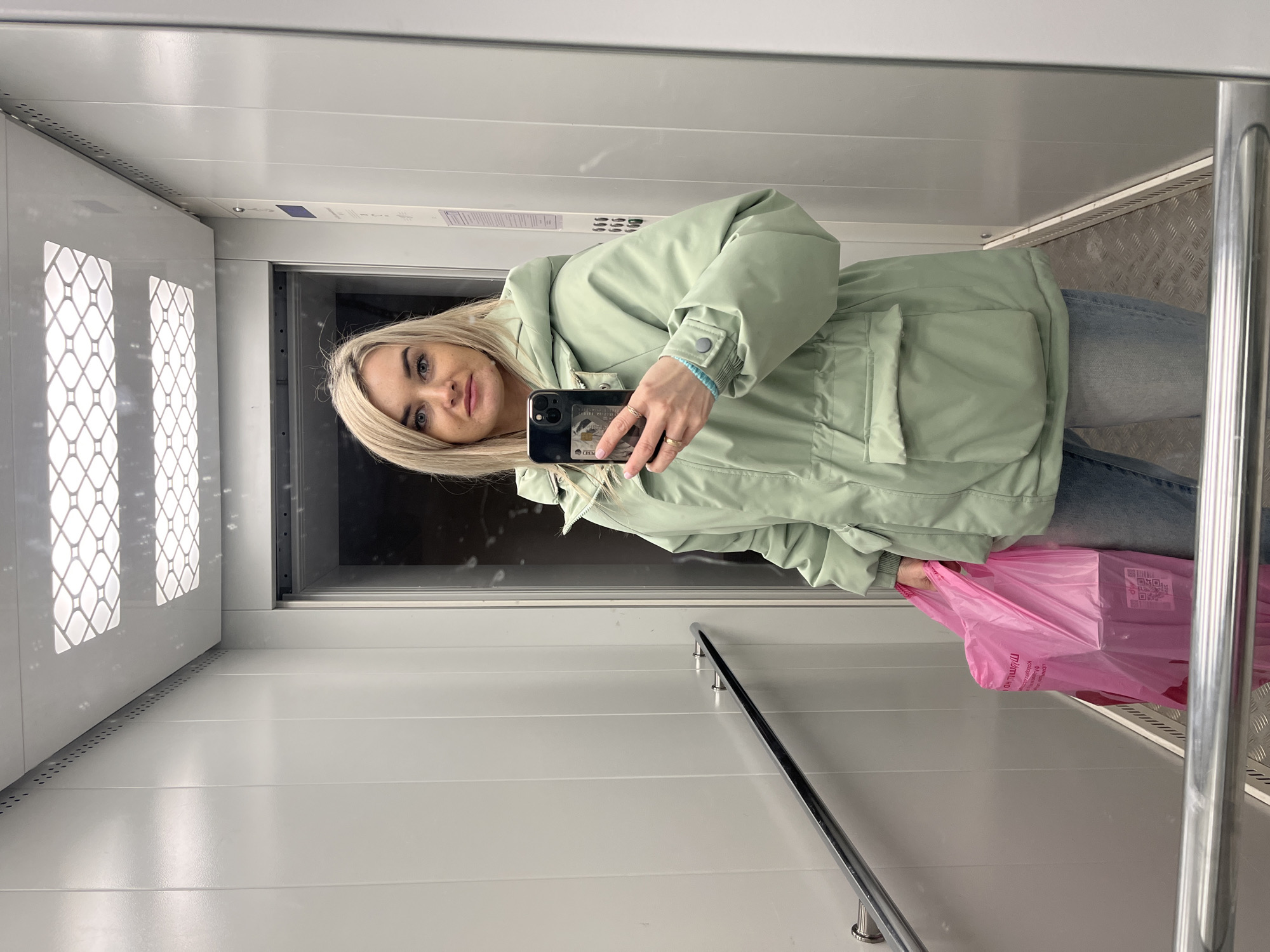 A young blonde woman is captured in a mirror selfie inside what appears to be an elevator or a small, reflective room. The image is taken at eye level, showing her from approximately the waist up. She has light-colored eyes and a slight smile, holding a black smartphone with a decorative case in her right hand to take the picture. Her left hand holds a vibrant pink plastic bag.
She is wearing a light mint green or sage-colored winter jacket with large pockets and a drawstring waist, paired with light blue jeans. Several rings are visible on her fingers. A light blue beaded bracelet is on her right wrist.
The setting consists of white or light gray walls, one of which is highly reflective, acting as the mirror for the selfie. Behind her to the left are elevator doors with a distinctive pattern of white square and diamond shapes, likely made of translucent material. Above these doors, on the wall or ceiling, is a small blue display panel, possibly an elevator floor indicator. To the right of the frame, a polished metal handrail runs horizontally along the wall. The floor, partially visible at the bottom right, appears to be made of textured metal plates, often found in elevators or industrial settings, while other parts show flat white panels.
The pink plastic bag she holds has some text and a QR code printed on it. The text includes "Минимар" (Minimar) followed by partially obscured words and "KORONA", suggesting a minimart or shop named "Minimart Korona" located in Chaikovsky, Russia. The woman appears to be in a pleasant, neutral mood, engaged in taking the self-portrait.