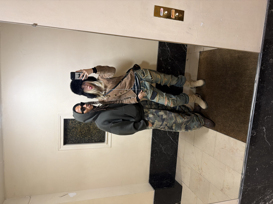 The image captures a mirror selfie of two individuals, reflected in what appears to be a large mirror in an indoor setting, likely an elevator lobby or hallway. The image is oriented sideways, with the subjects appearing to stand vertically on the right side of the frame as if they are standing on a wall.
The person in the foreground (closer to the camera's perspective within the reflection) is on the left side of the frame. This individual appears to be male, wearing a dark grey or black hooded sweatshirt and camouflage-patterned pants. They have dark dreadlocks or braided hair, and are wearing dark rectangular sunglasses. One hand is partially visible, possibly in a pocket or resting at their side. They are wearing dark shoes or boots.
Behind them, slightly to the right in the reflection, is the second person, who appears to be female. She is holding a black smartphone up to her face, taking the mirror selfie. She has long blonde hair, partially covered by a black, furry hat. She is wearing a light brown or tan-colored jacket with multiple buttons down the front, possibly over a striped top. Her pants are blue jeans with significant rips, distressing, and patches. She is wearing light-colored, possibly off-white, boots. Her tongue is slightly sticking out, giving a playful expression.
The background visible in the reflection includes light-colored, possibly beige or off-white walls. On the wall behind the second person's head, in the upper right portion of the reflected image, there is a golden-colored panel with two circular buttons, typical of an elevator call panel. Above the top button, the word "LOBBY" is clearly visible. Below the bottom button, there is some faint, unreadable text.
The floor in the reflection is a mix of light-colored tiles, possibly marble, and darker, possibly black, sections or tiles. A dark brown mat or rug is visible on the floor in the lower right section of the reflection. To the left of the male figure, on the wall, there is a small, rectangular recessed area or panel, possibly a window or decorative element, with a patterned texture visible inside.
The overall impression is a casual, fashion-forward mirror selfie taken by two individuals, possibly waiting for an elevator or in a building lobby.