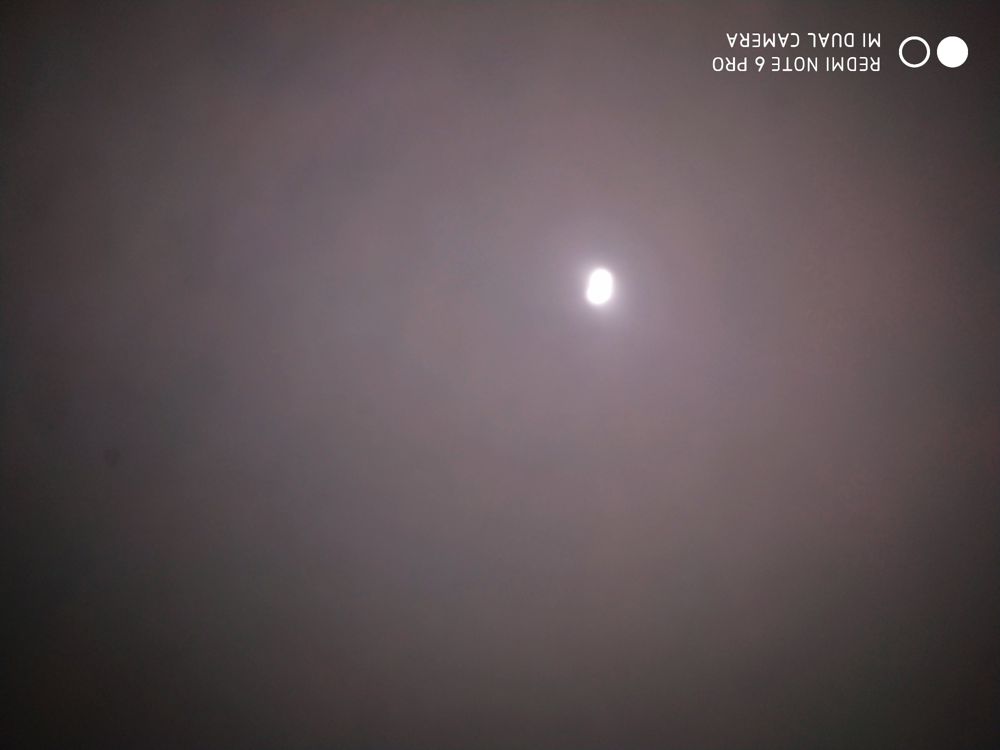 The image shows a hazy, overcast sky with a bright, circular object, possibly the sun or moon, partially obscured by the atmospheric conditions. The object is located in the upper left quadrant of the image. A watermark in the lower left corner indicates "REDMI NOTE 6 PRO MI DUAL CAMERA". The overall impression is of a celestial body seen through a veil of mist or fog. There are no visible people, buildings, or specific geographical landmarks that would allow for precise location identification within Badas, Indonesia.