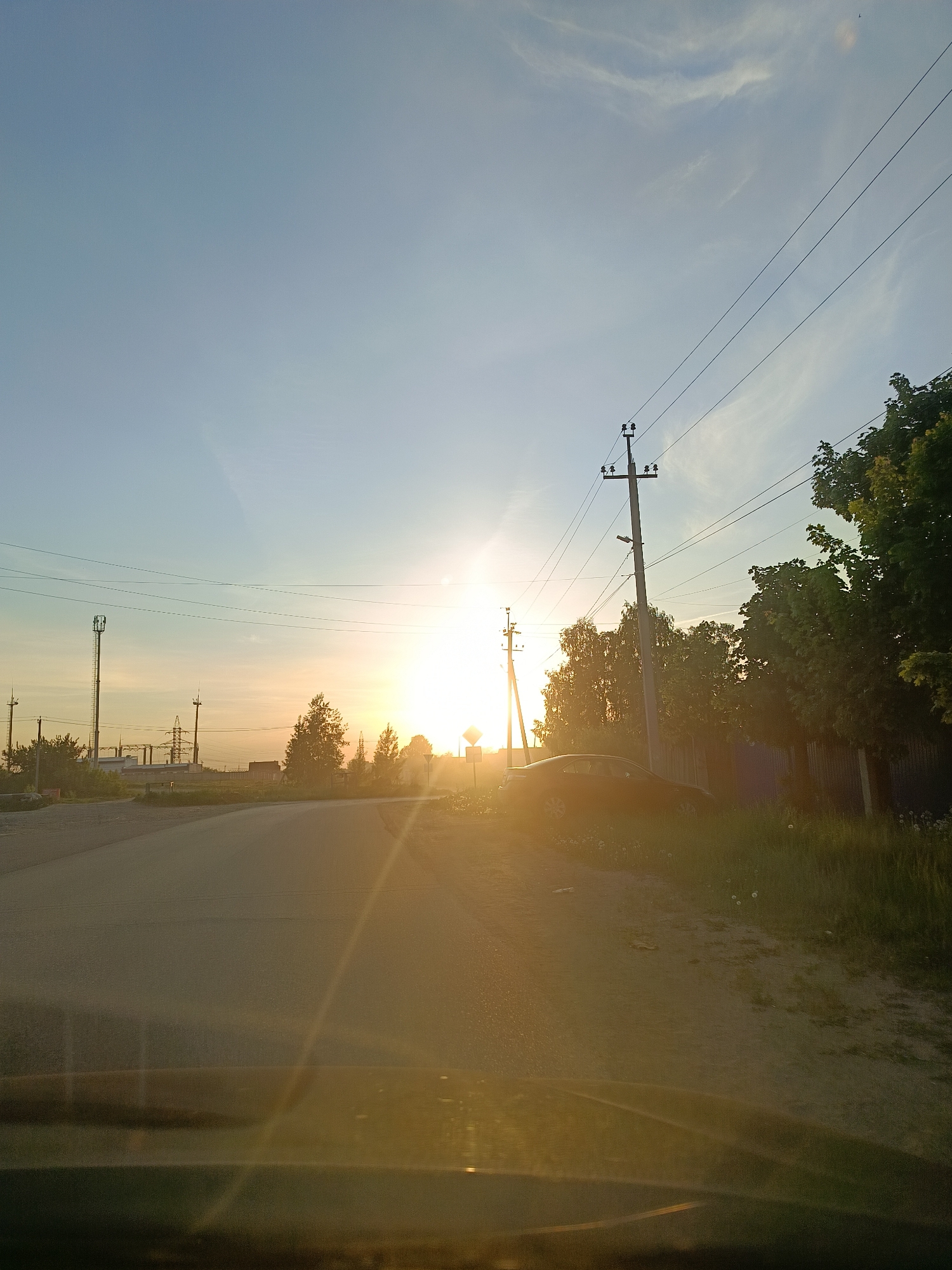 The image depicts a road scene in Chaikovsky, Russia, during either sunrise or sunset. The sun is positioned centrally, close to the horizon, creating a strong glare. The road stretches into the distance, with a slight curve. On the right side of the road, there are power lines and poles, a fence, trees, and a parked dark-colored car. In the background, on the left side of the road, there's an industrial area with communication towers and what appears to be a substation. The image appears to have been taken from inside a vehicle, with the dashboard visible at the bottom.