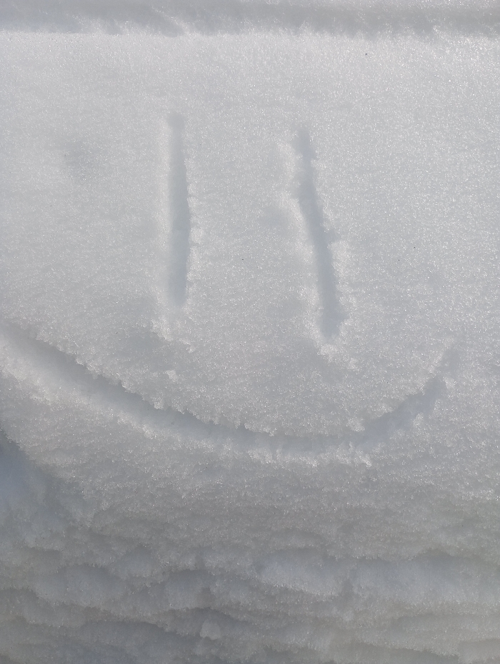 The image displays a close-up, downward-angled view of a snow-covered surface in the city of Яромаска, Russia. The snow is white and granular, exhibiting varied texture and subtle shadows from natural light. Etched into the upper-middle section of the snow is a simplified smiley face, formed by three distinct, shallow indentations: two parallel vertical lines serving as eyes, and a single upward-curving line beneath them acting as a mouth. The carvings are precise, creating clear depressions against the slightly raised snow surface. The lower portion of the image reveals more pronounced undulations and layering within the snow, suggesting depth or accumulation. No individuals, animals, or man-made objects other than the snow etching are present in the scene. The overall environment indicates outdoor winter conditions.