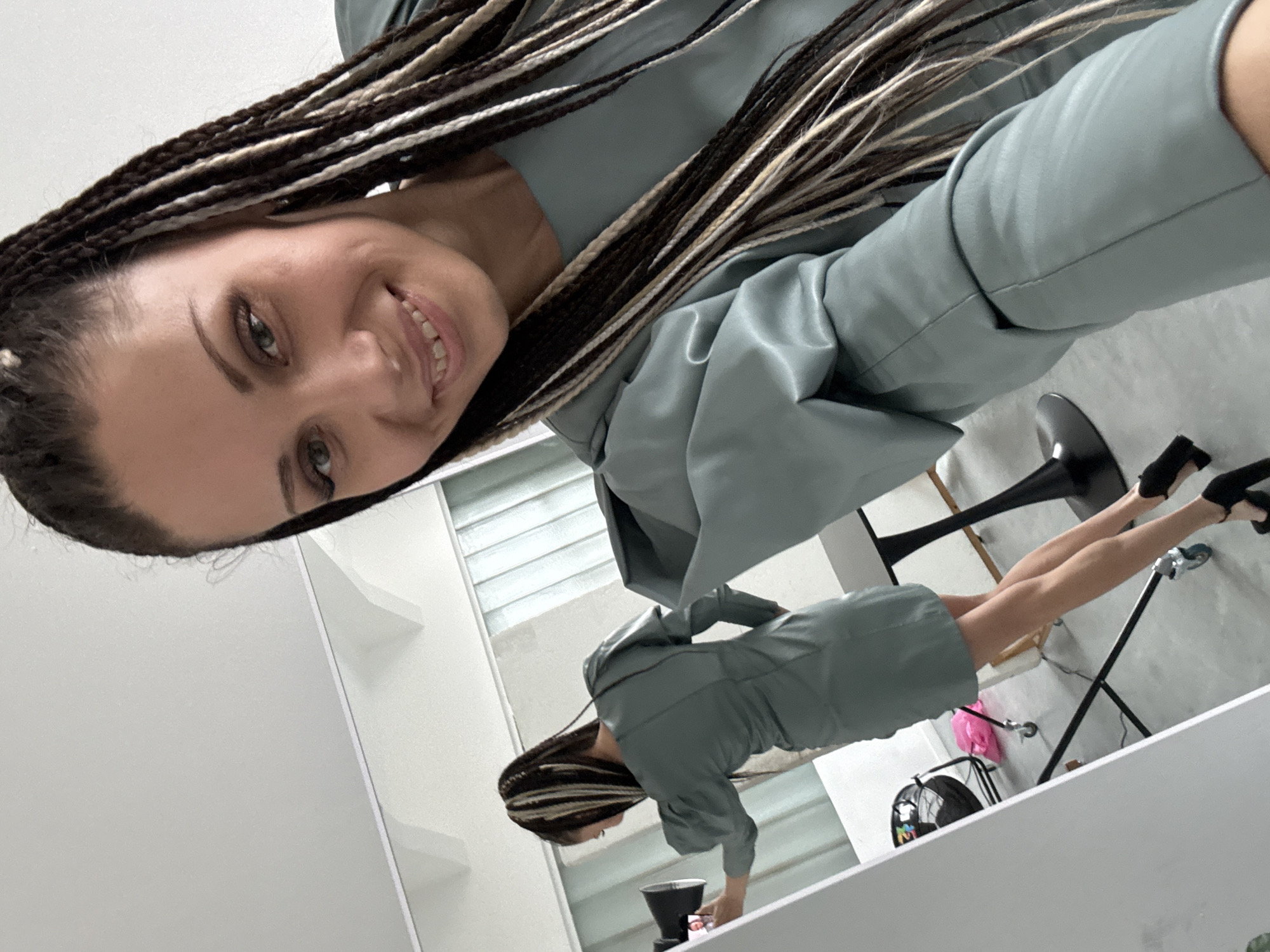 A fair-skinned woman with braided hair (predominantly dark with some white strands) poses for a selfie indoors, in what appears to be a dressing area or studio. She is wearing a blue-gray colored outfit, possibly a dress or a jacket with a dress. A mirror reflects the woman's full-length image standing by a white counter, appearing to touch something out of view. There is a black backpack visible in the reflection, along with some lighting equipment, including a stand with a pink bag attached to it. She is wearing black heels. A black chair with a black base is visible in the foreground. There is no identifiable information to determine the location as Bor, Russia.