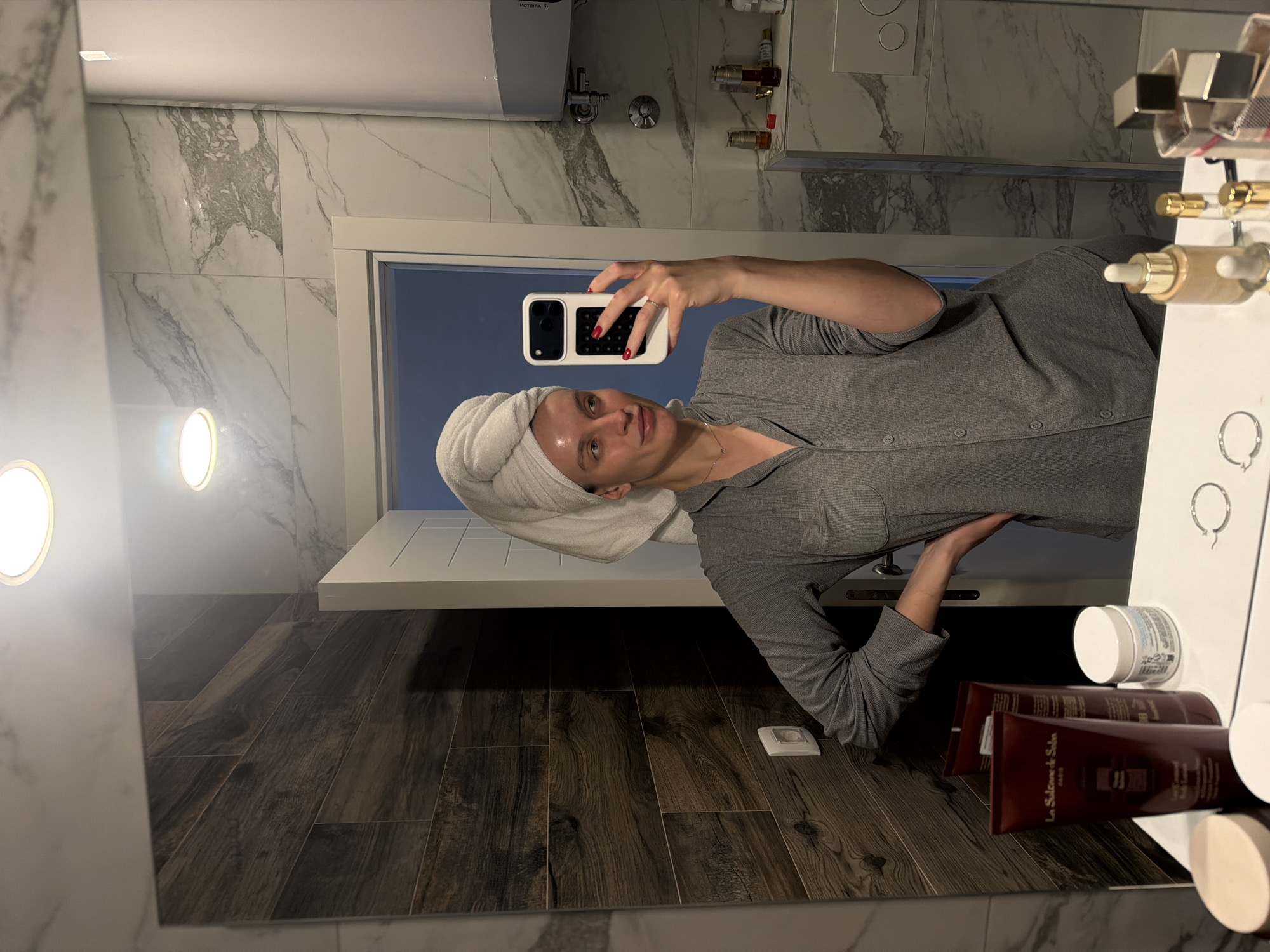 A woman with a towel wrapped around her head is taking a selfie in a bathroom. She is wearing a grey button-up shirt. The bathroom has grey and white marble walls and dark brown wood-look flooring. There is a white sink counter to her right, with various skincare products and accessories. A white-framed doorway is behind her. The location is in Budva, Montenegro.