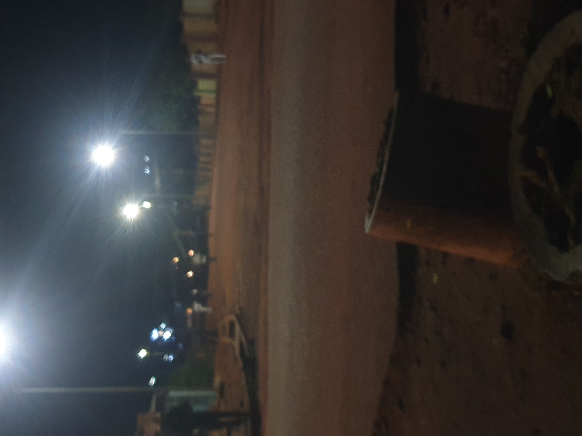 This is a nighttime, slightly out-of-focus photograph taken on a dirt road in the city of Jos, Nigeria. Several streetlights illuminate the scene, casting a bright glow and creating starburst effects. In the background, there are a few indistinct figures of people walking or standing. To the left, a person in dark clothing is partially visible. The road surface appears to be made of reddish-brown dirt. In the foreground, a cylindrical object, possibly a pipe or a concrete form, is positioned upright. Further in the foreground, a dark object sits on the ground, with some lighter elements suggesting it might be food or debris on a surface. The overall environment appears to be an outdoor, possibly residential or public space within Jos.