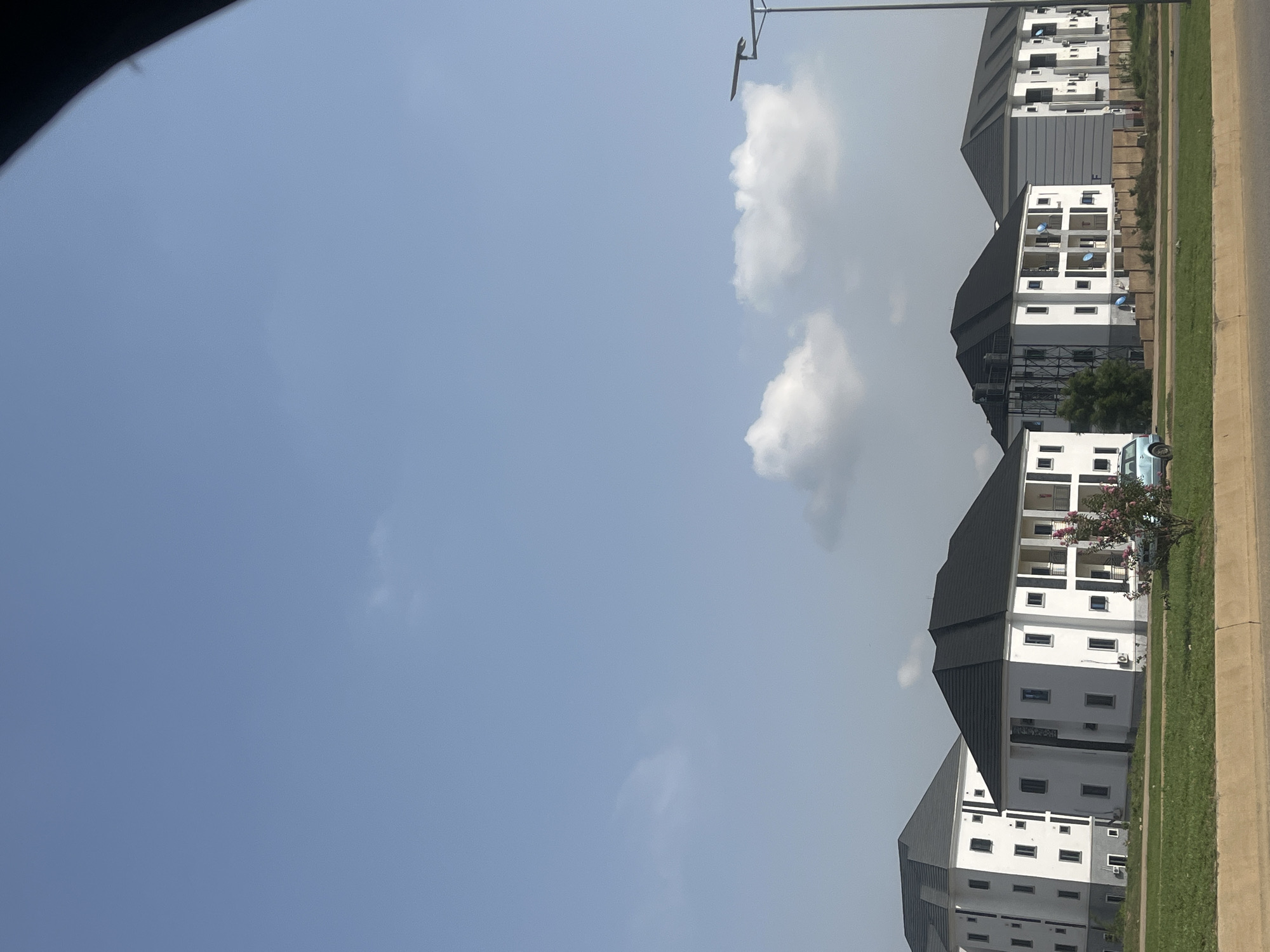 This image depicts a row of modern, multi-story residential buildings with white facades and dark tiled roofs, situated in the city of Karum, Nigeria. The buildings appear to be recently constructed or undergoing finishing touches, as evidenced by the presence of various architectural features like balconies and multiple windows. A light blue vehicle is parked in front of one of the buildings. The foreground shows a well-maintained green lawn bordered by a concrete barrier, suggesting a planned residential development. The sky is clear with a few scattered white clouds. A streetlamp is visible on the right side of the frame. The overall scene suggests a quiet, possibly suburban, residential area.