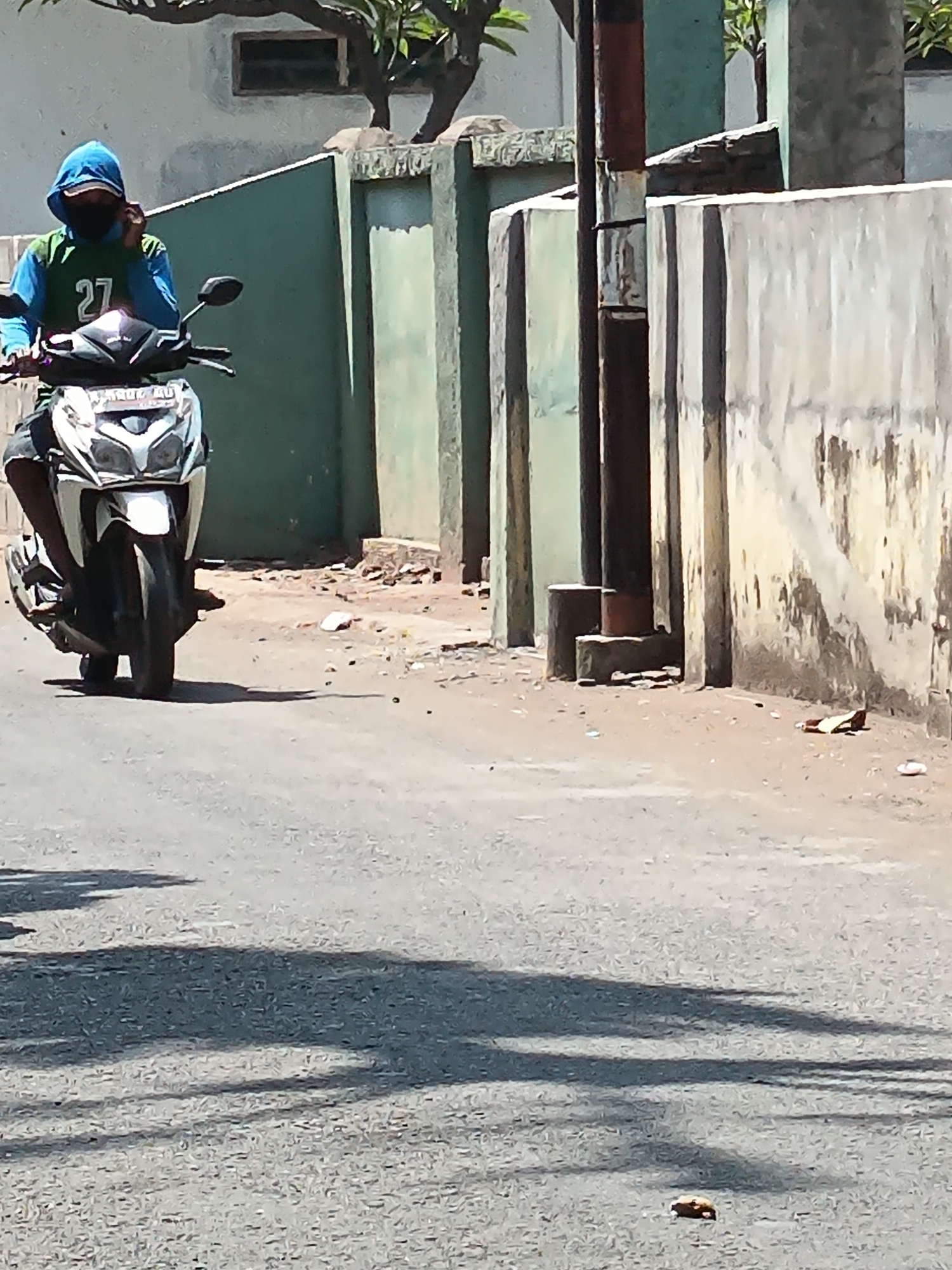 A person wearing a blue hoodie and a face mask rides a white scooter down a paved road in Kandangjati Wetan, Indonesia. The scooter has a license plate and the rider is wearing a green and blue shirt with the number "27" on it. The background consists of a textured wall and a green wall, with some foliage and trees visible. The road is lined with a concrete barrier. Shadows of trees are cast across the road. There is some debris on the side of the road.