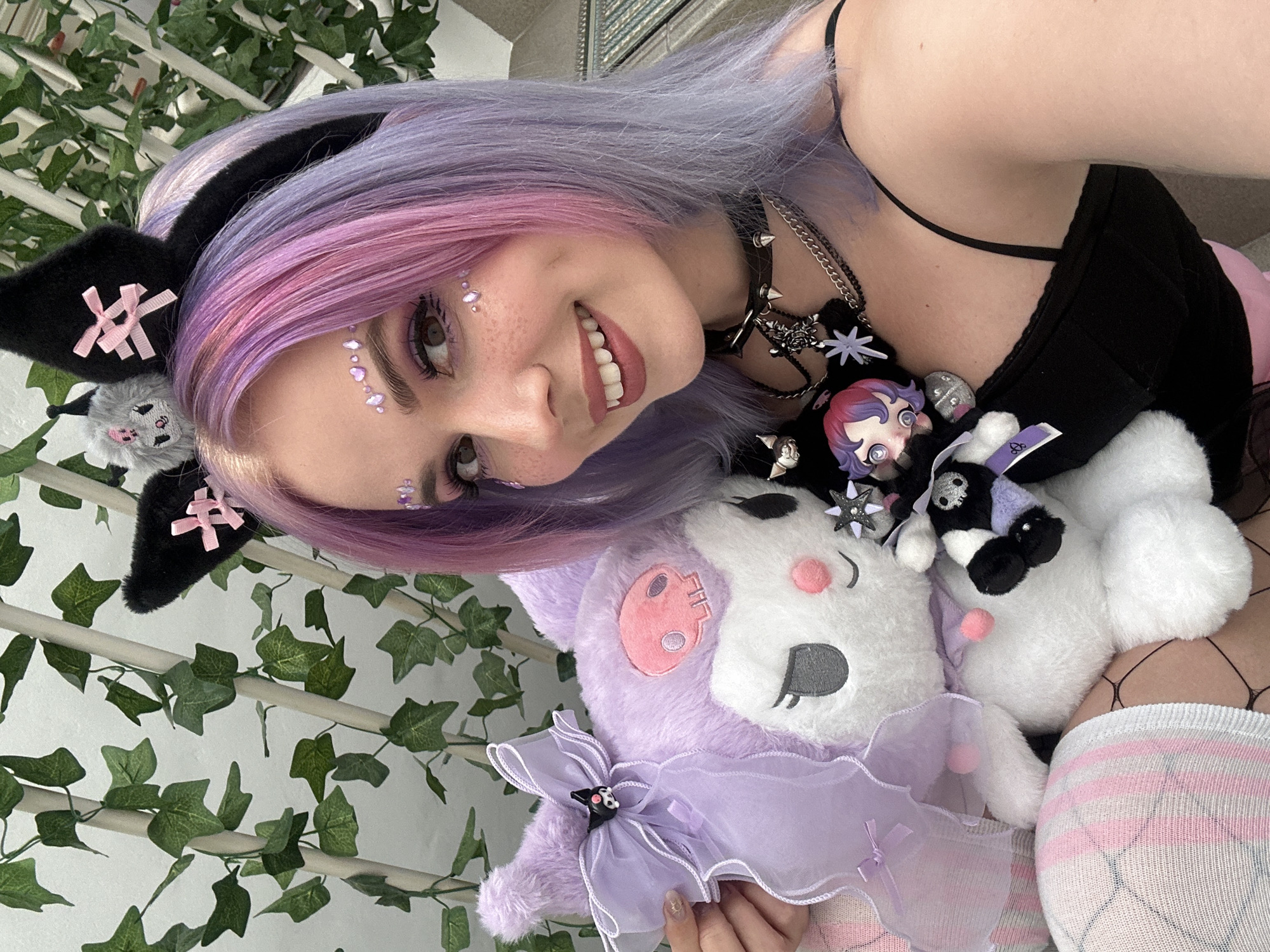 A young woman with purple and pink hair, wearing cat ears and sparkly makeup, is holding several plush toys, including one that is lavender-colored with a sheer ruffle around its neck. She is also wearing a black choker with spikes and several necklaces. The background includes artificial ivy. There is no verifiable evidence to suggest that this photograph was taken in the city of Zagreb, Croatia.
