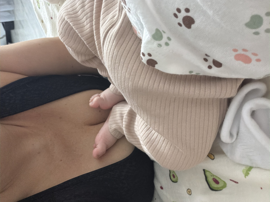 This image captures a close-up, indoor scene in Chaikovsky, Russia, depicting a partial view of an adult and an infant. The upper chest and left shoulder of an adult are visible on the left side of the frame, showing light-toned skin and a black garment with a wide strap, revealing the cleavage area. An infant's lower body, including legs and bare feet, is positioned on the right, with the infant's feet resting in contact with the adult's chest. The infant is dressed in light brown, vertically ribbed fabric pants. The immediate environment includes layered textiles: a white fabric with a repeating pattern of green avocado halves and smaller yellow/orange shapes is visible on the lower left, and another white fabric featuring brown, light green, and pink paw prints is visible on the lower right. A small, folded white textile item is also present at the bottom center of the frame.