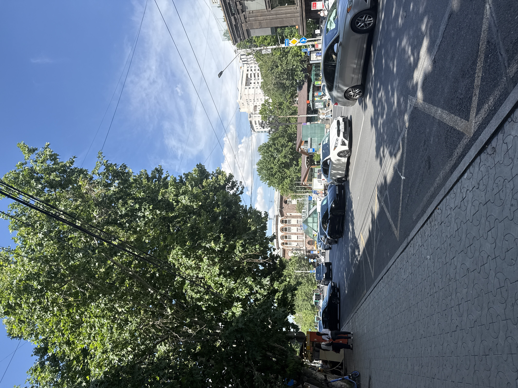 The image depicts a sunny day in a city. The scene is a street with cars parked and driving on the right side of the road. A sidewalk with a patterned paving stones runs along the right side of the street. There are trees on the left side of the street, with their branches casting shadows on the road. Buildings are visible in the background.
Several people are visible on the sidewalk, some near a small vendor stall. The time of day appears to be daytime, given the bright sunlight and shadows. The sky is blue with scattered white clouds. The presence of power lines and streetlights suggests an urban environment. The architecture of the buildings hints at a location in Yerevan, Armenia. No specific event or activity is clearly discernible beyond the general context of a city street scene. There is no visible text in the image.
