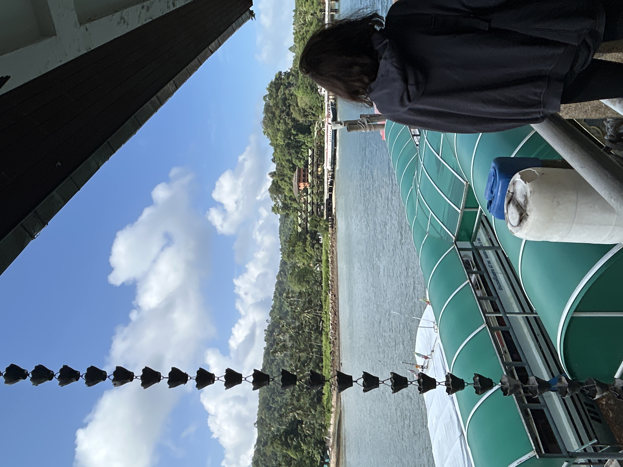 A person wearing a dark hooded sweatshirt stands on a pier, facing away from the camera, looking out at a body of water. In the foreground, a chain of black, flower-shaped gutter cups hangs vertically. Beyond the pier, several boats with green canopies are docked. The water is calm, reflecting the blue sky with scattered white clouds. Lush green hills covered in trees and vegetation rise from the opposite shore. On the distant shore, buildings with terracotta roofs and a pier are visible. The scene is likely the Pak Nam district of Krabi province, Thailand, given the tropical island scenery and piers that are common there.