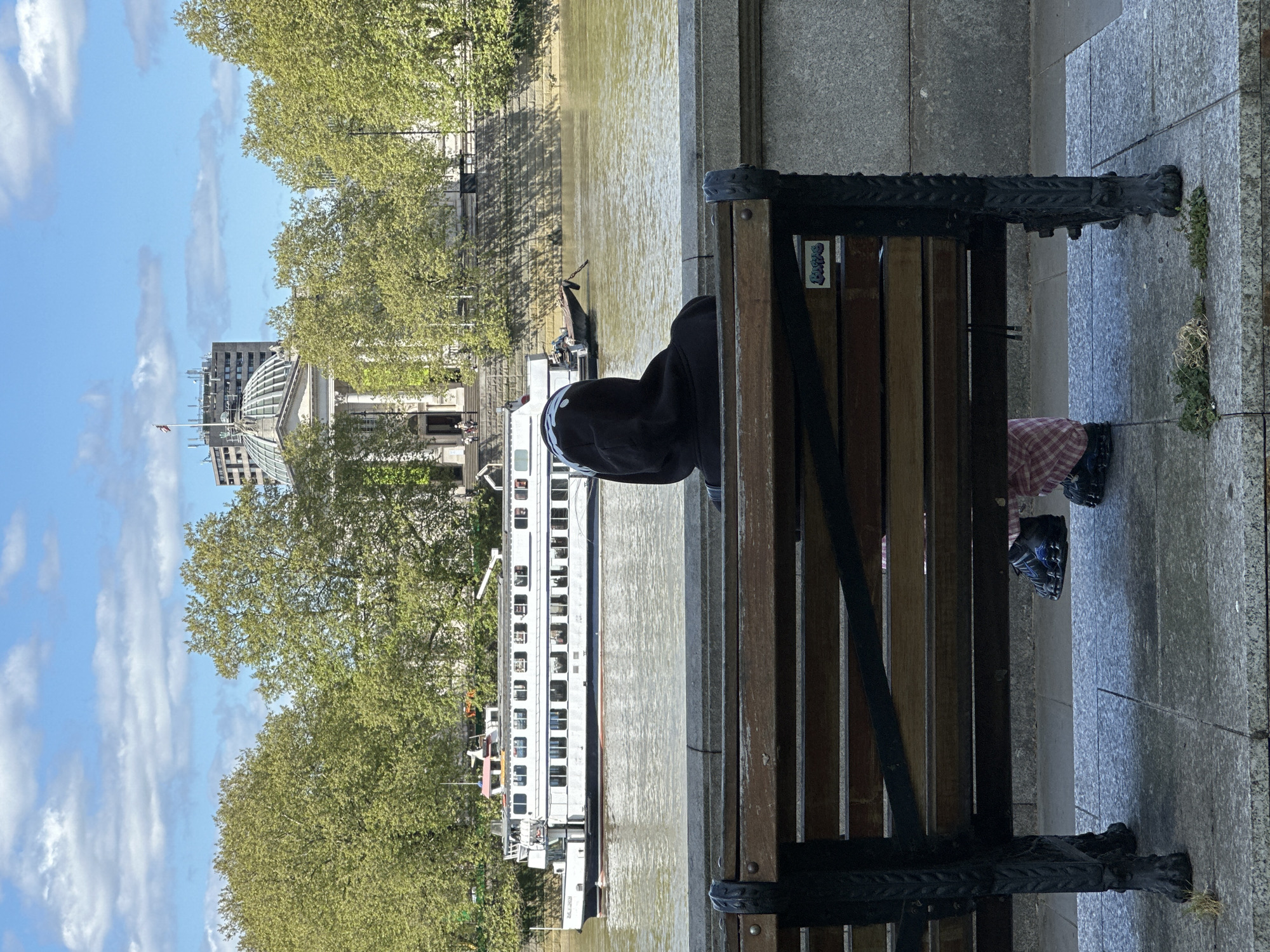 A person sits on a park bench next to the River Thames in London, United Kingdom. The person wears dark clothing and their face is obscured by a hood. The bench is made of wood and metal, and is placed on a stone path beside a stone wall. The river reflects the sunlight. A white boat floats on the river. Trees line the opposite bank of the river, and a building with a domed roof is visible in the background. The sky is blue with white clouds.