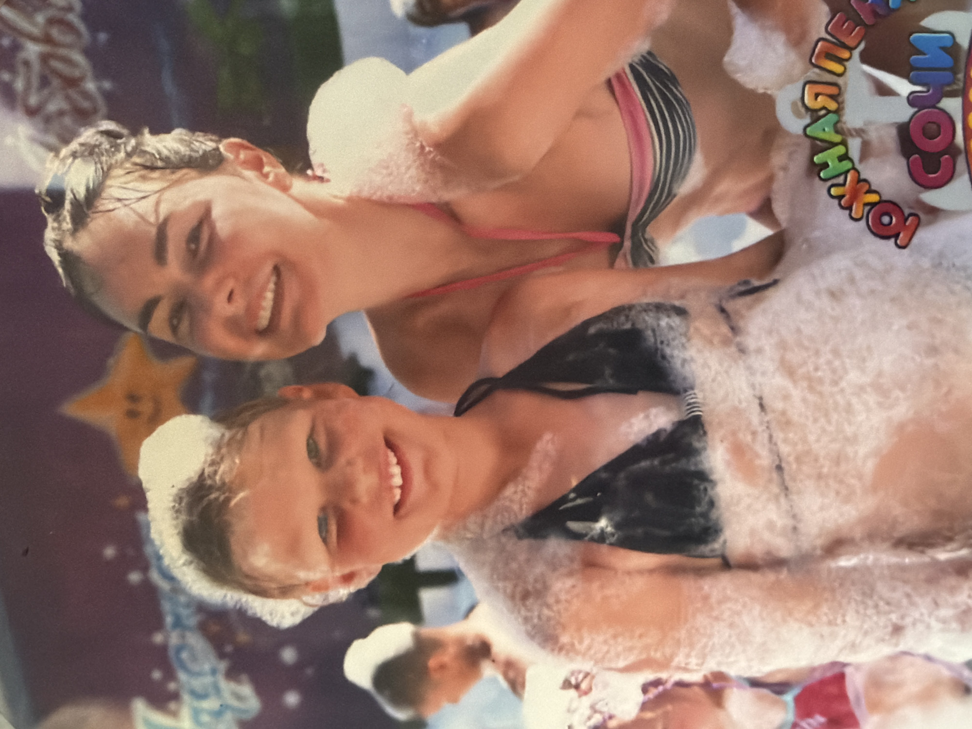 Two young women in bathing suits are covered in white foam. The woman on top is wearing a striped bikini top with a pink strap, while the woman below has on a dark bikini top. Both are smiling. A sign with the words "СОЧИ ПЕРВАЯ" in colorful lettering is partially visible near the top right. The location appears to be Sochi, Russia, as indicated by the sign. Other blurred figures can be seen in the background.