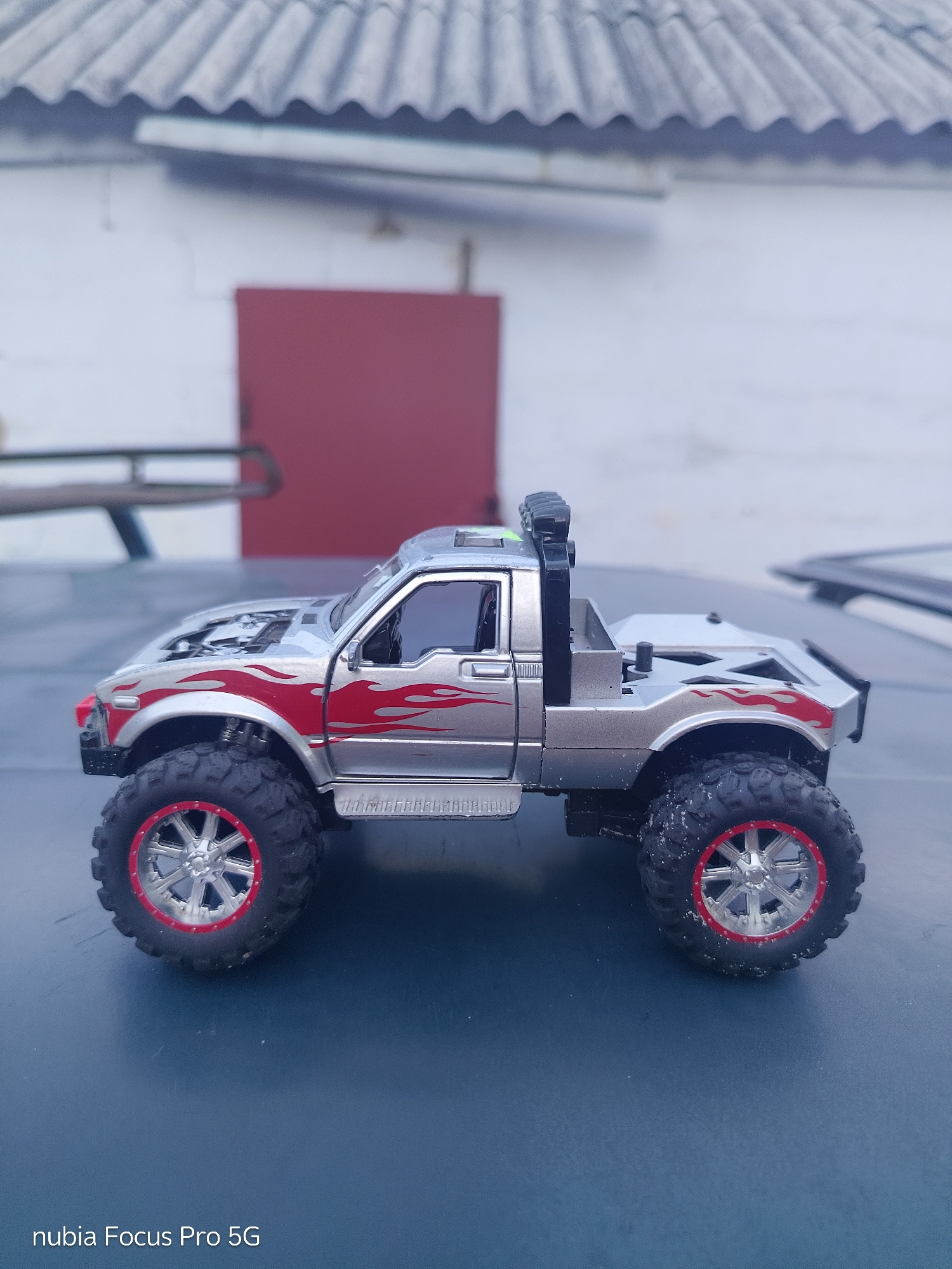 A silver toy monster truck with red flame decals and red-accented wheels is parked on a dark blue surface. The truck is positioned in profile view, facing left. The background consists of a white wall with a red door and a corrugated roof. The scene appears to be outdoors, possibly in an urban or suburban setting. The overall impression is a still life photograph of a toy.