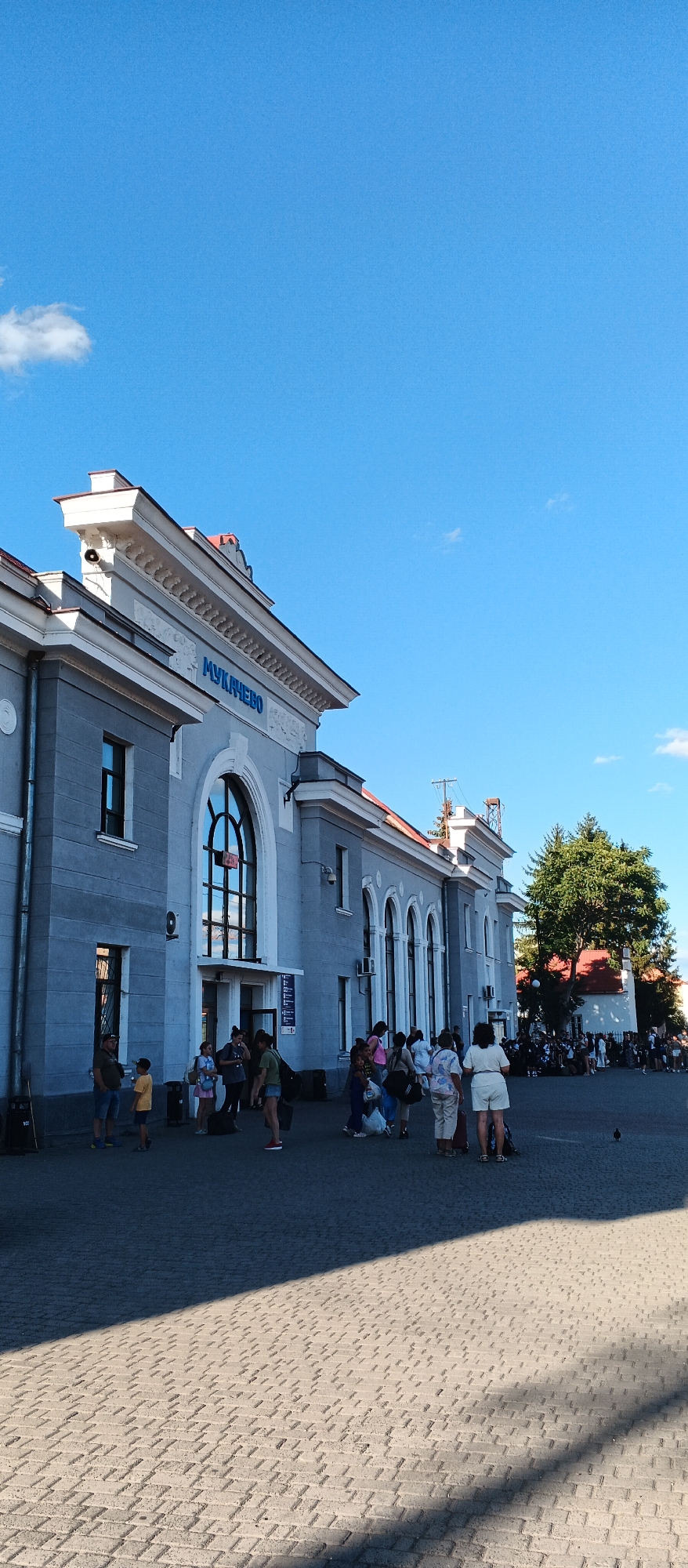 The image depicts the Mukachevo Railway Station in Ukraine on a clear, sunny day. The station building is a light grey, multi-story structure with a classical architectural style, featuring arched windows and decorative cornices. The name "МУКАНЧЕВО" is visible in blue lettering above the main entrance.
In front of the station, a paved plaza is filled with people. A diverse group, including adults and children, are seen standing, walking, and carrying luggage, suggesting they are either arriving or departing. Some individuals are interacting with each other, while others appear to be waiting. The ground is paved with interlocking bricks, and a strong shadow from an unseen object bisects the plaza.
In the background, a large green tree stands to the right of the station, and further behind it, another building with a red roof is partially visible. The sky above is a bright, clear blue with a few wispy white clouds. A lone pigeon is observed on the paved ground in the lower right quadrant of the image. The overall scene conveys a typical day at a busy transportation hub.
