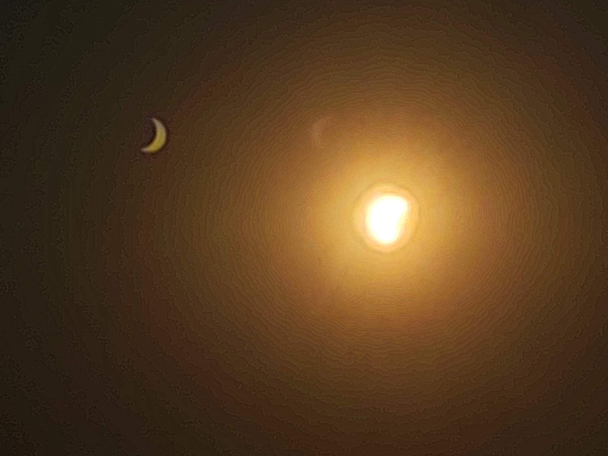 A crescent moon is visible in the upper left quadrant of the image, against a dark, hazy sky. The moon is illuminated and appears yellow, with a distinct curved shape. To the right of the moon, and occupying the majority of the image, is a bright, diffuse light source, likely the sun, appearing as a glowing orb with a yellowish-orange hue. The sky around the light source exhibits a gradient of light intensity, with brighter tones near the source and gradually darkening towards the edges of the image. Subtle circular patterns or textures are visible within the illuminated areas of the sky, possibly indicative of atmospheric conditions or optical effects. The overall scene suggests a celestial event, such as a solar eclipse, occurring at dusk or dawn, or a photograph taken with a strong light source that has caused lens flare. The specific location in Autillo de Campos, Spain, is not visually determinable from the image itself.