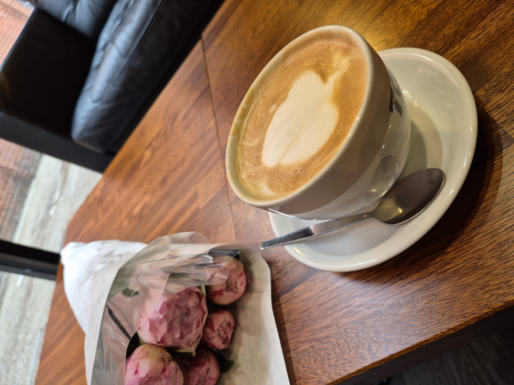 This image captures an intimate indoor setting, likely in West Suffolk, United Kingdom, showcasing a cup of coffee and a small bouquet of flowers on a polished wooden table. The main focus is a white ceramic coffee cup filled with a latte, featuring clear white latte art, resembling a heart, on its frothed surface. A reflective silver spoon rests on the matching white saucer.
Adjacent to the coffee, a delicate bouquet of pink flowers, appearing to be peonies, is bundled in plain white paper and encased in clear plastic wrapping. Several full blooms are visible, contributing a soft, natural element to the scene. These items are positioned on a rich, reddish-brown wooden table, whose distinct grain and polished surface are illuminated by ambient indoor light, possibly enhanced by natural light from a nearby window.
In the upper left background, part of a dark, presumably black, leather sofa with characteristic tufted detailing is visible, suggesting a comfortable seating arrangement. Beyond the sofa, a blurry area hints at an exterior view or an adjacent space, possibly through a window. The overall impression is one of quiet contemplation or a pleasant break. No discernible text is present within the image.
