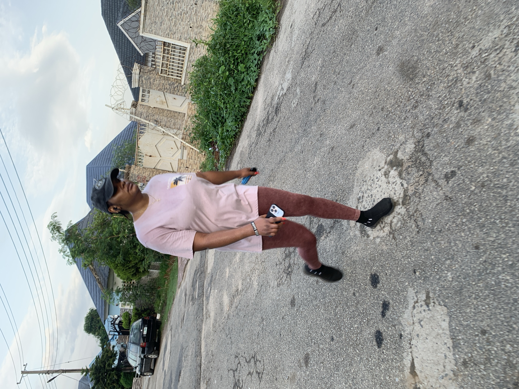 A woman wearing a pink t-shirt, brown leggings, black socks, and black shoes is walking on a gray paved road. She is holding a mobile phone in her right hand. She wears a black cap. The background includes houses, greenery, and overhead electrical wires. The location appears to be a residential street in Abuja, Nigeria. A car is visible parked along the side of the road. The time of day appears to be late afternoon.